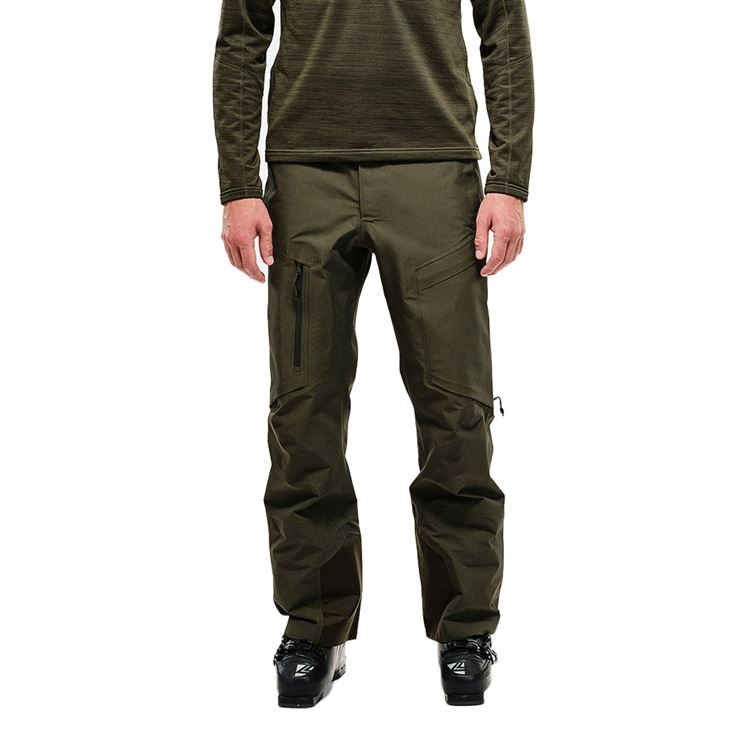 Mann in Skikleidung, trägt eine grüne Y-1 HD GORE-TEX Pro 3L Shell Pant und Skischuhe. Hose mit Taschen und Reißverschlüssen.