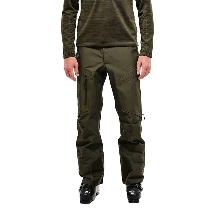 Uomo con abbigliamento da sci, indossa un pantalone Y-1 HD GORE-TEX Pro 3L Shell verde e scarponi da sci. Pantaloni con tasche e cerniere.