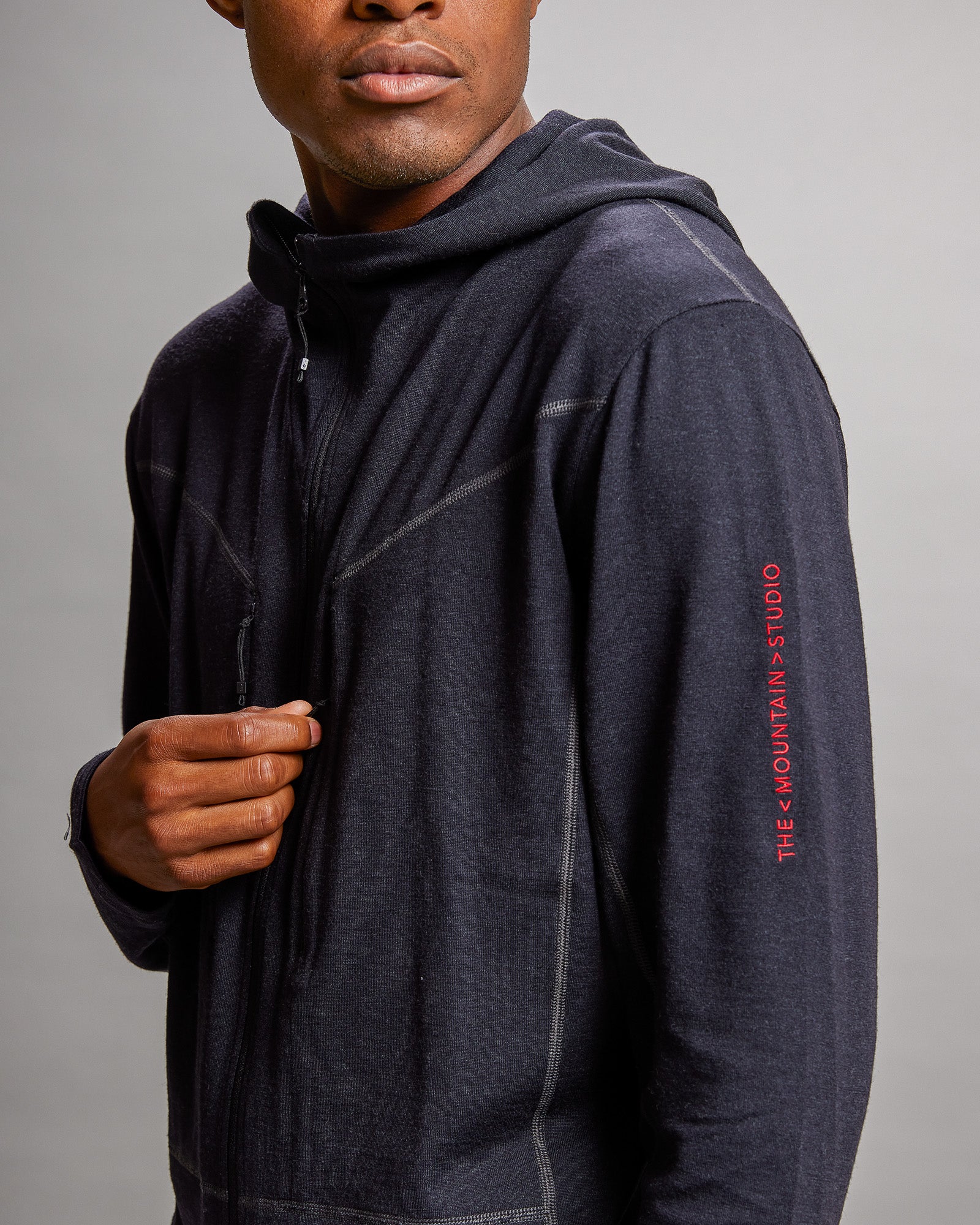 Nahaufnahme einer schwarzen NUYARN Hood Jacket mit Kapuze und Reißverschluss am Oberkörper eines Mannes. Rotes Logo am Ärmel.