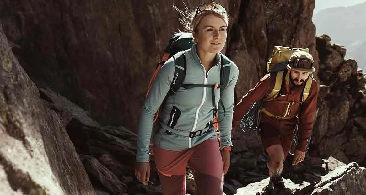 Two hikers climb up a path between rocks, the woman in gray leads, the man in red follows. Both are carrying rucksacks.