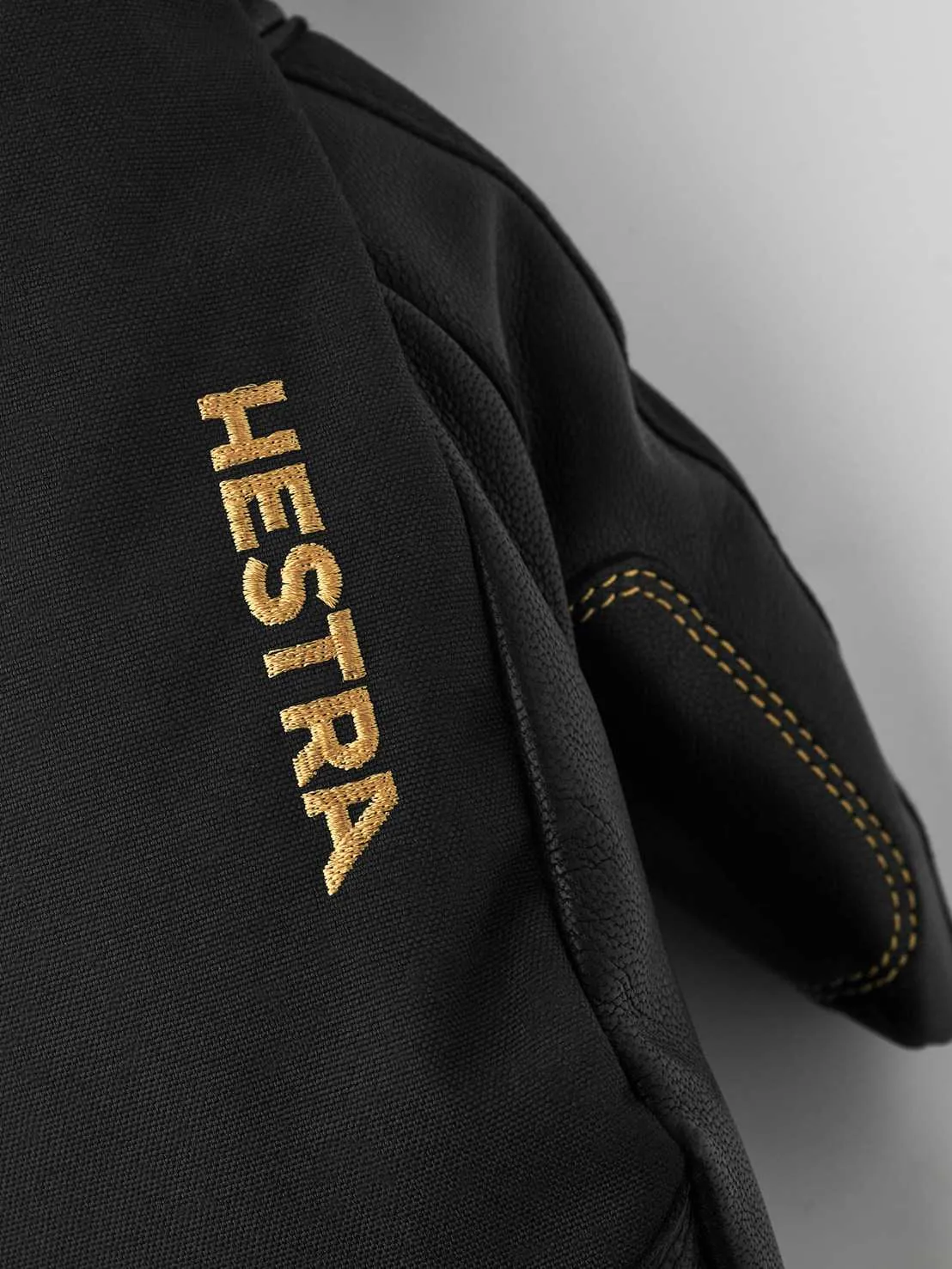Detailed view of a black Hestra glove with golden 'Hestra' embroidery and yellow stitching.