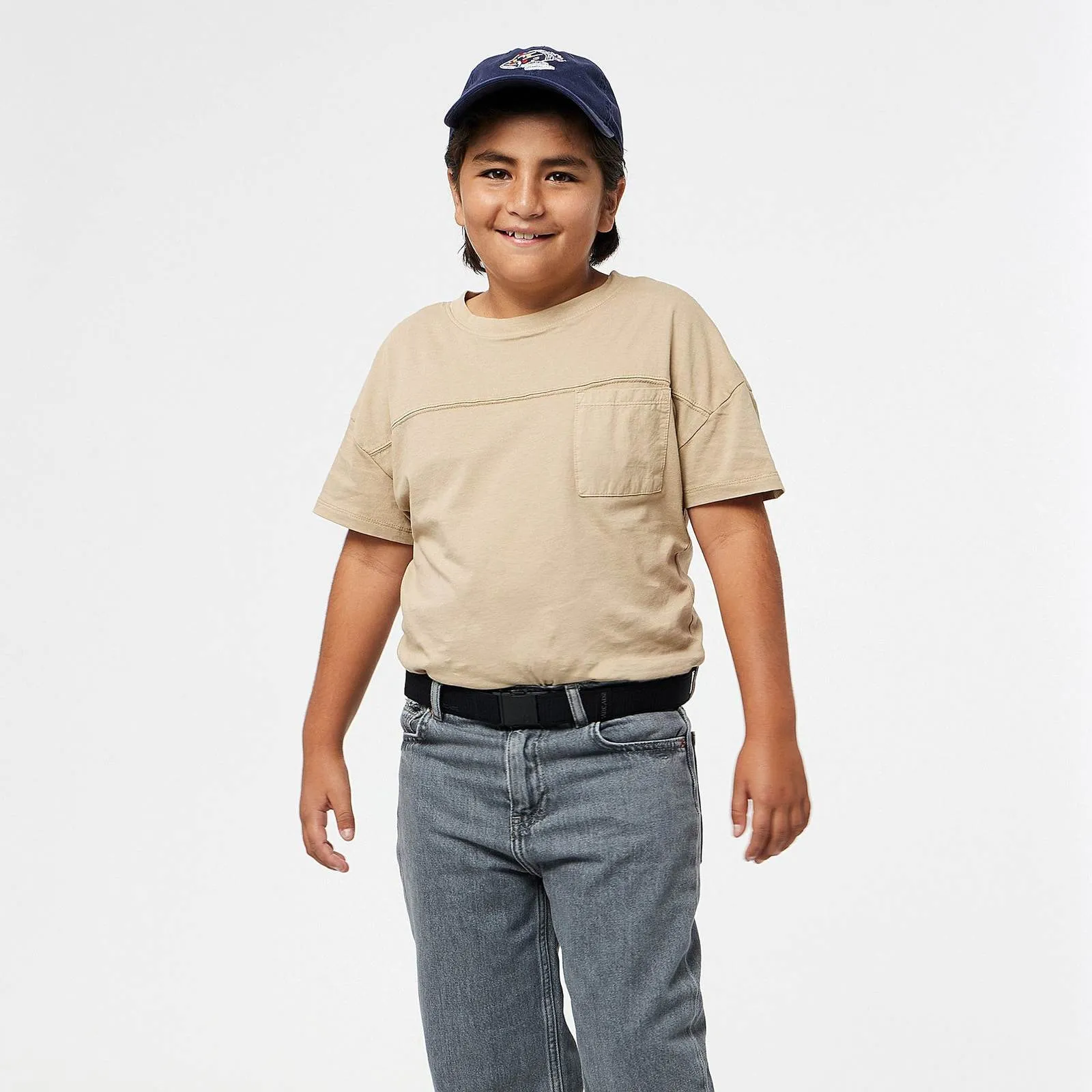 Enfant avec une casquette de baseball bleue et un T-shirt beige, portant une ceinture noire Arcade Atlas avec un jean gris.