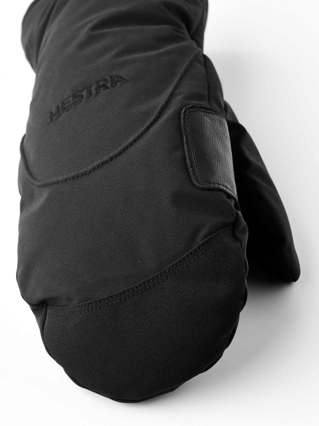 Gros plan sur une moufle noire avec un logo discret Hestra et un accent en cuir sur un fond clair.