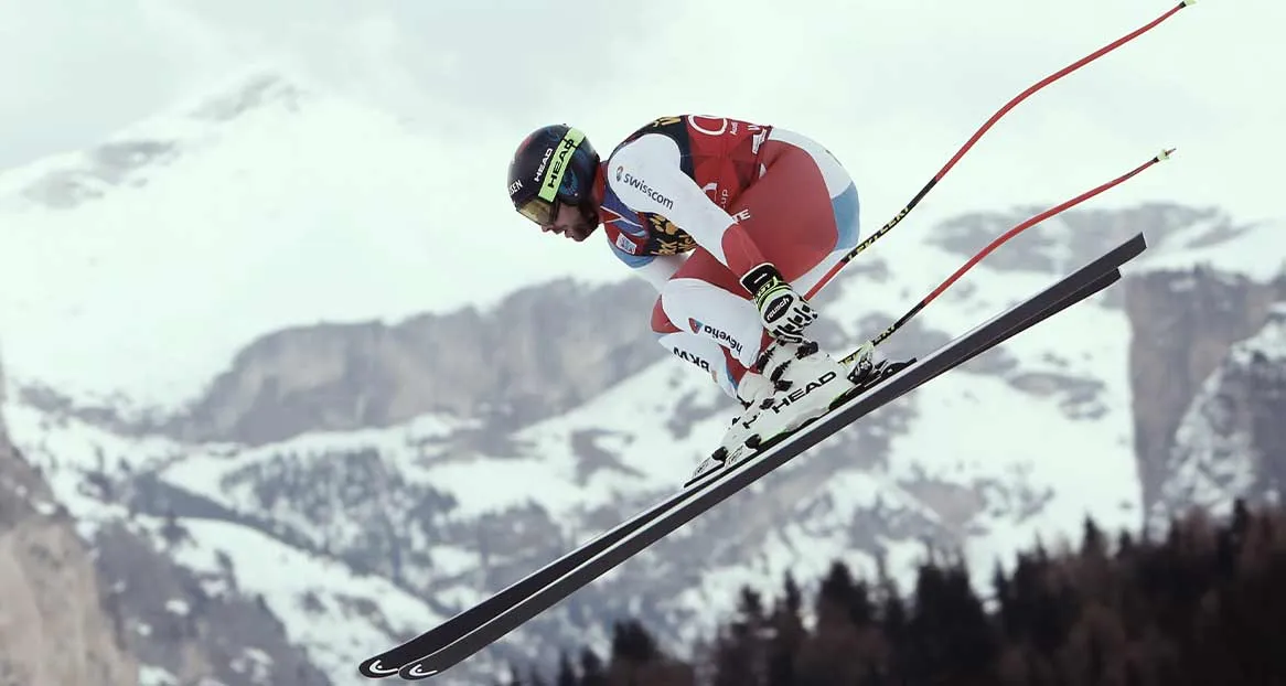 Ein Skirennläufer in roter und weißer Rennkleidung springt über eine Piste vor schneebedeckten Bergen.