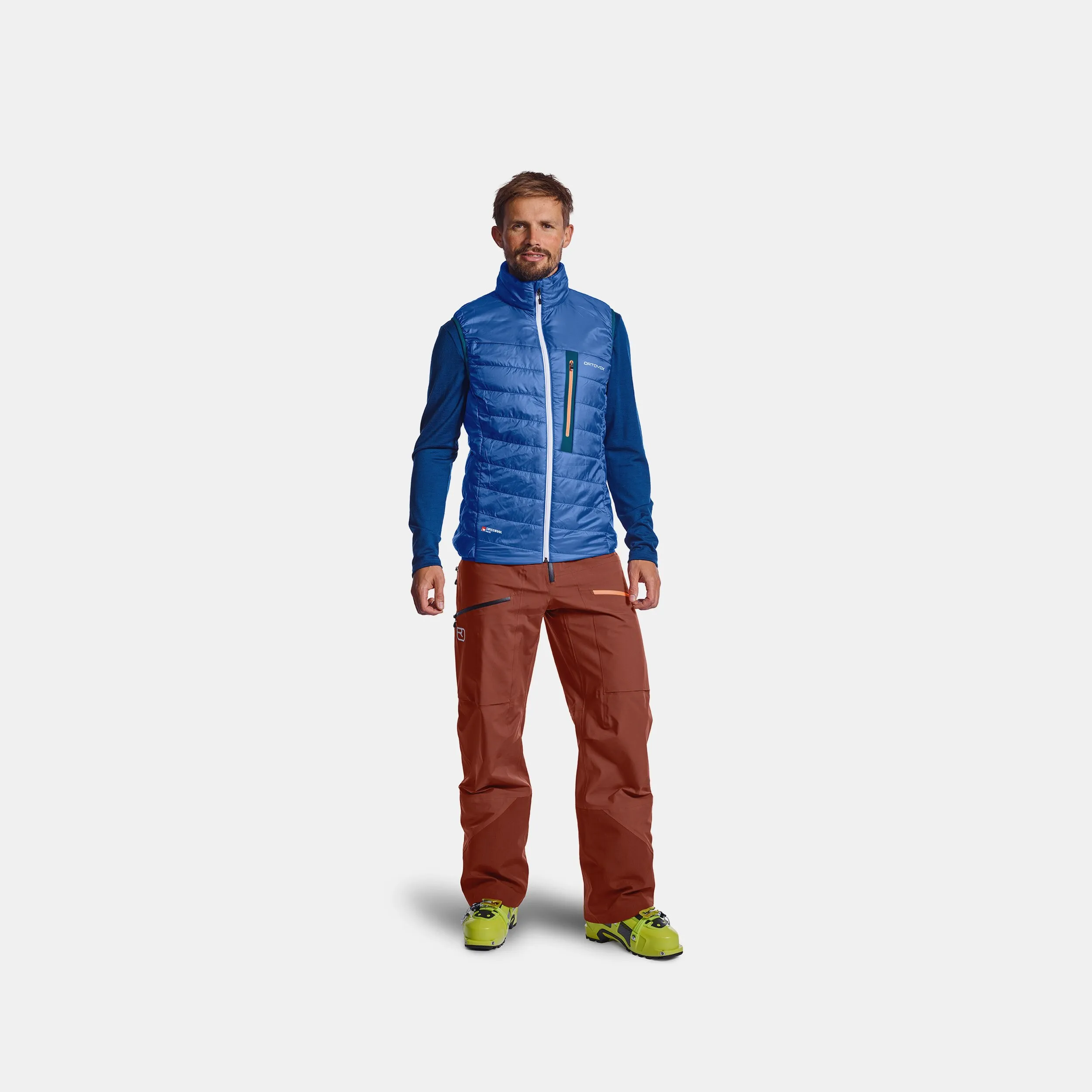 Man wearing blue Ortovox PIZ Cartas vest, red pants and ski boots against a neutral background.