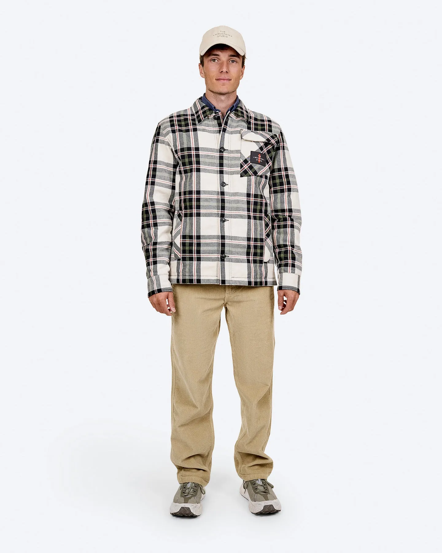 Ein Mann in Cordhose, karierter Jacke und Mütze steht vor einem weißen Hintergrund.