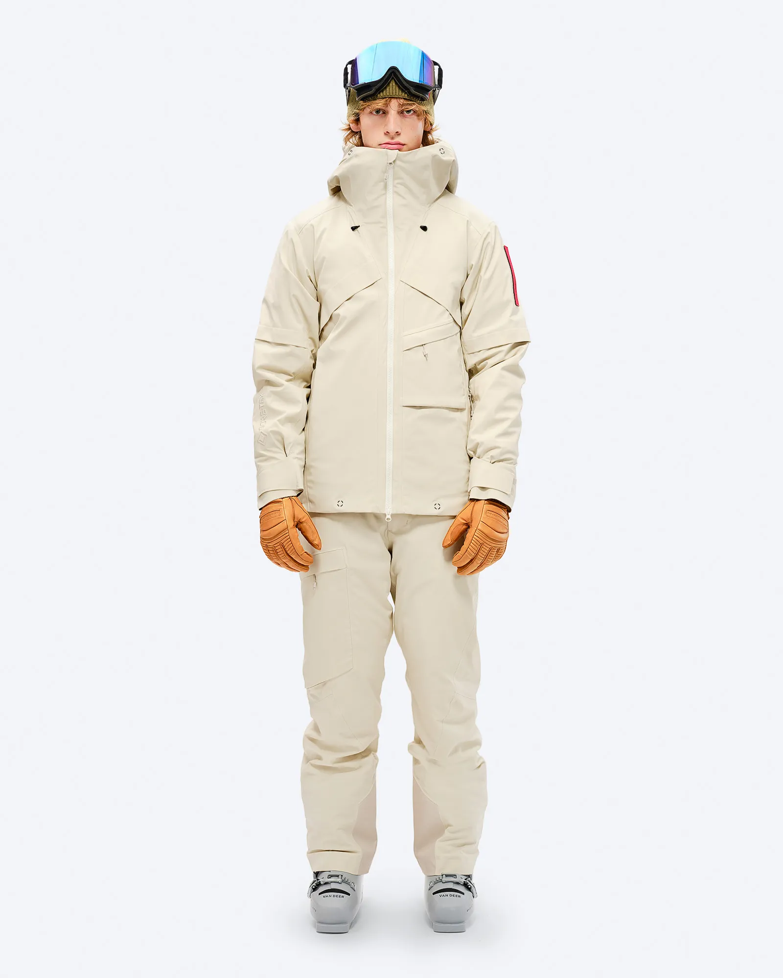 Person in beige ski/snowboard jacket with GORE-TEX, pants, gloves and ski goggles against a white background.