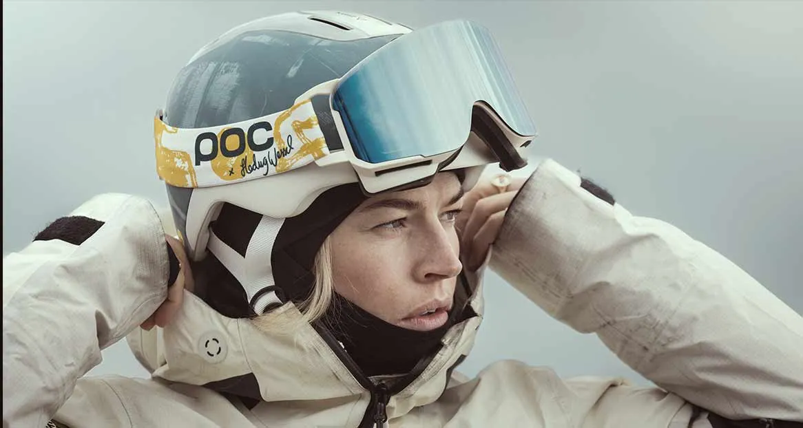 Woman with ski helmet and goggles, pulling on her jacket, looking to the side.
