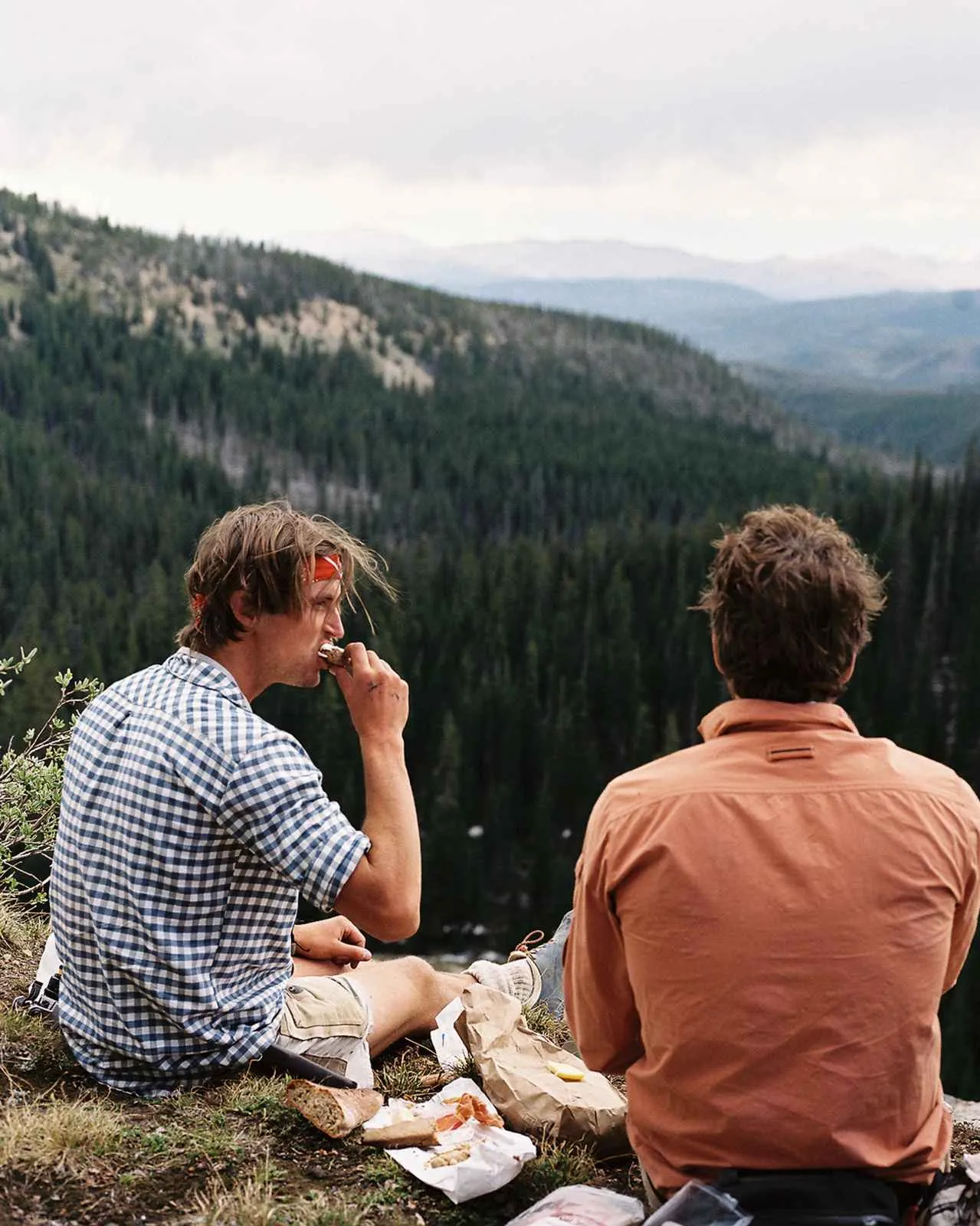 Two men are sitting on a hill overlooking a forest and mountains. One is eating, there is bread and provisions.