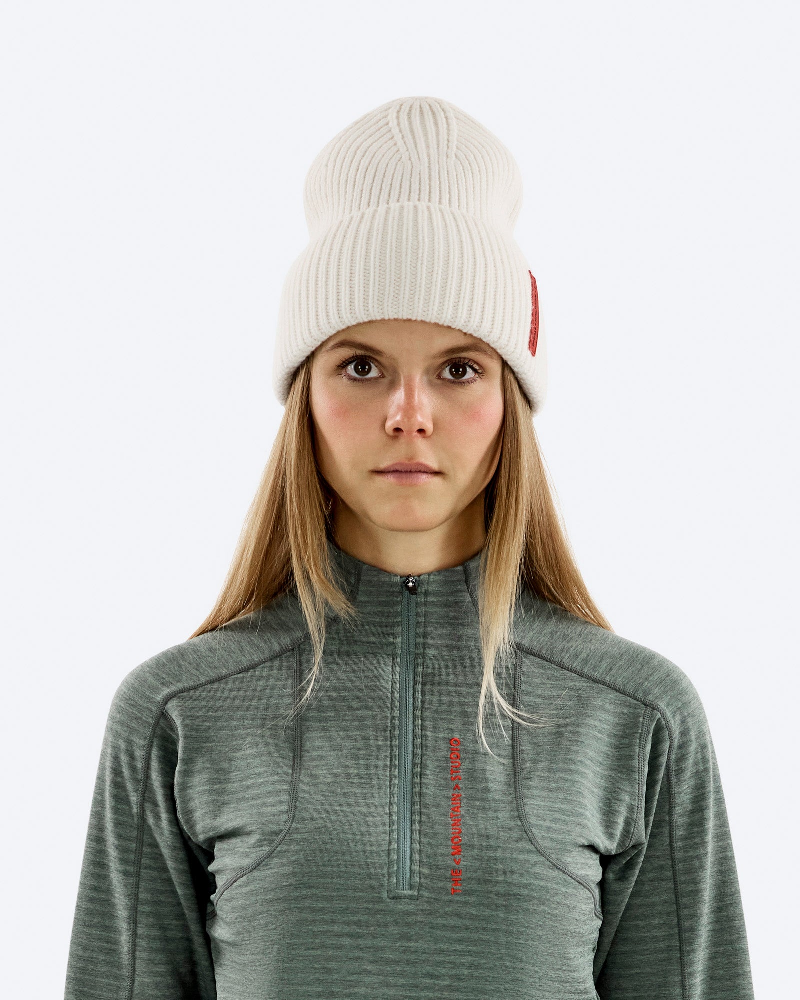 Porträt einer Frau mit blonden Haaren, die eine weiße Mütze und ein grünes Oberteil mit Reißverschluss trägt.