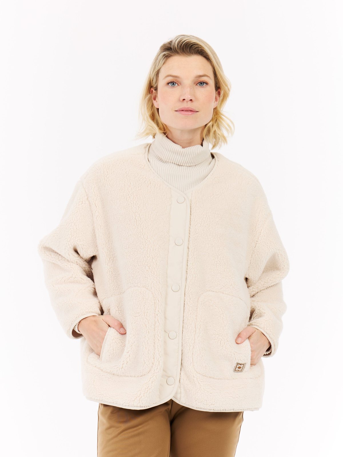 A woman with blonde, shoulder-length hair wears a cream-colored fleece jacket over a turtleneck sweater and brown pants. Her hands are in the pockets of the jacket.