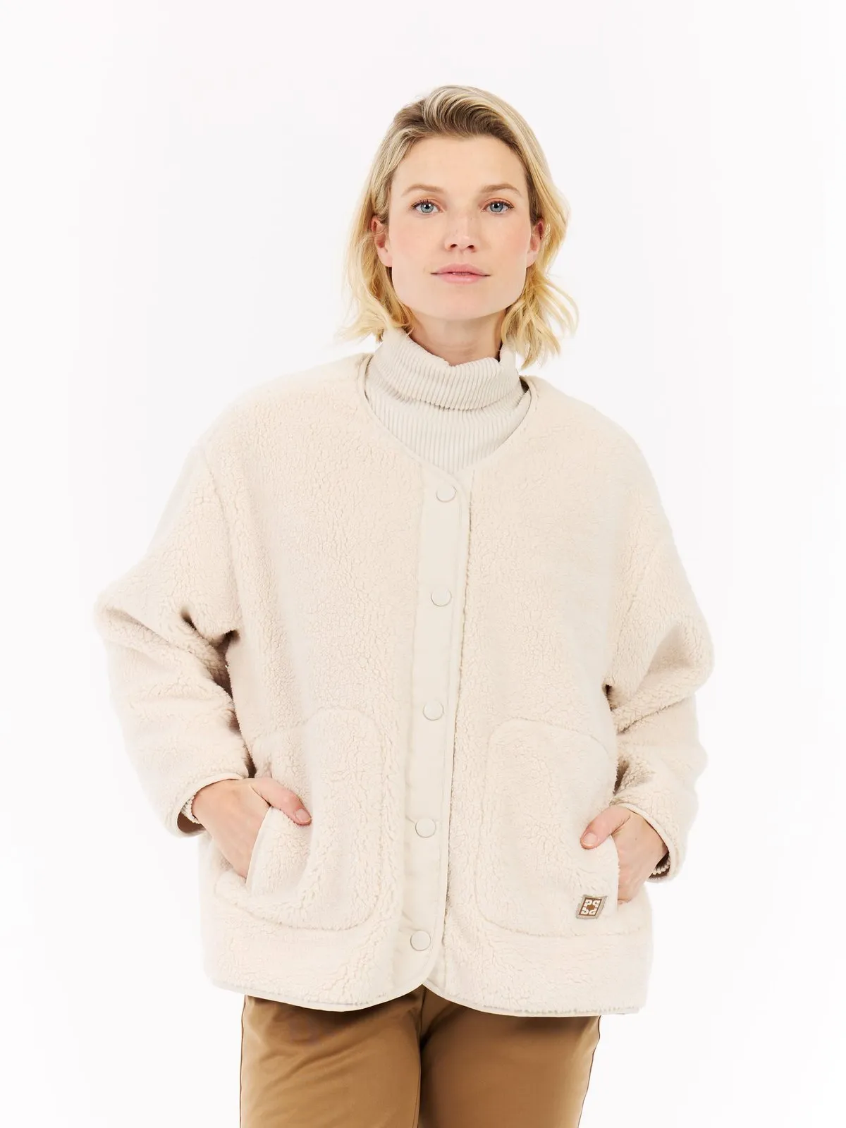 Eine Frau mit blonden, schulterlangen Haaren trägt eine cremefarbene Fleecejacke über einem Rollkragenpullover und braune Hosen. Ihre Hände stecken in den Jackentaschen.