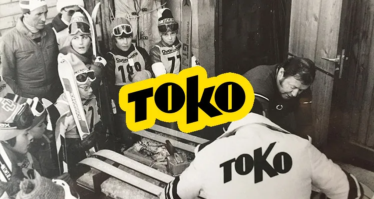 Schwarz-Weiß-Aufnahme einer Gruppe Skifahrer, einige Kinder mit Skibrillen und Rennanzügen, und zwei Erwachsene, die Skier präparieren, mit dem "TOKO"-Logo überlagert.