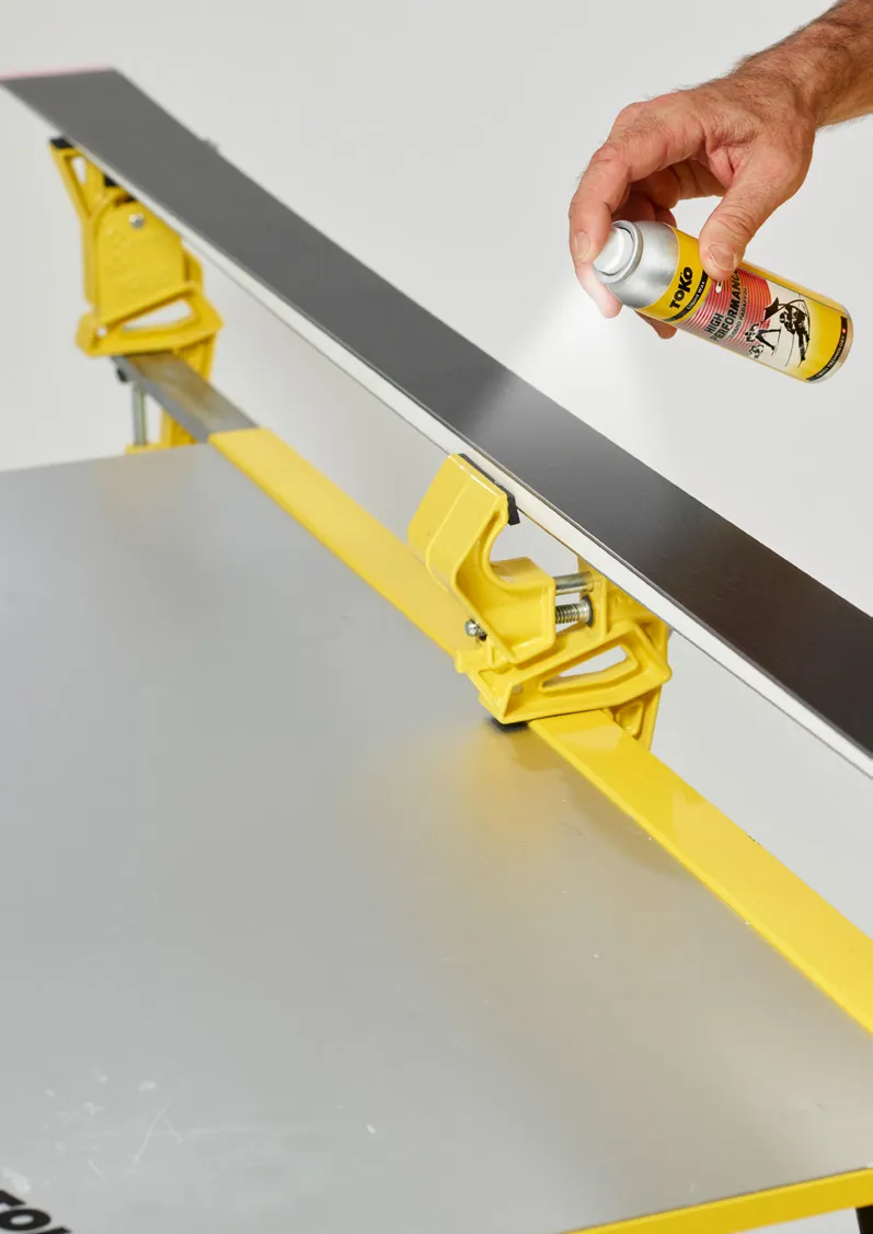 Une main vaporise du fart sur le dessous d'un ski fixé dans un établi jaune.