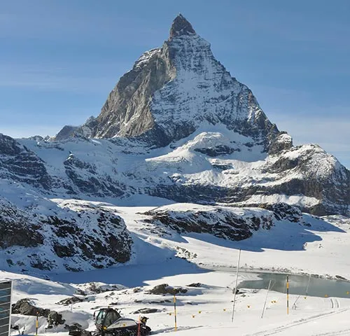 Una montagna innevata con una cima appuntita, circondata da un paesaggio invernale con un piccolo lago in primo piano.