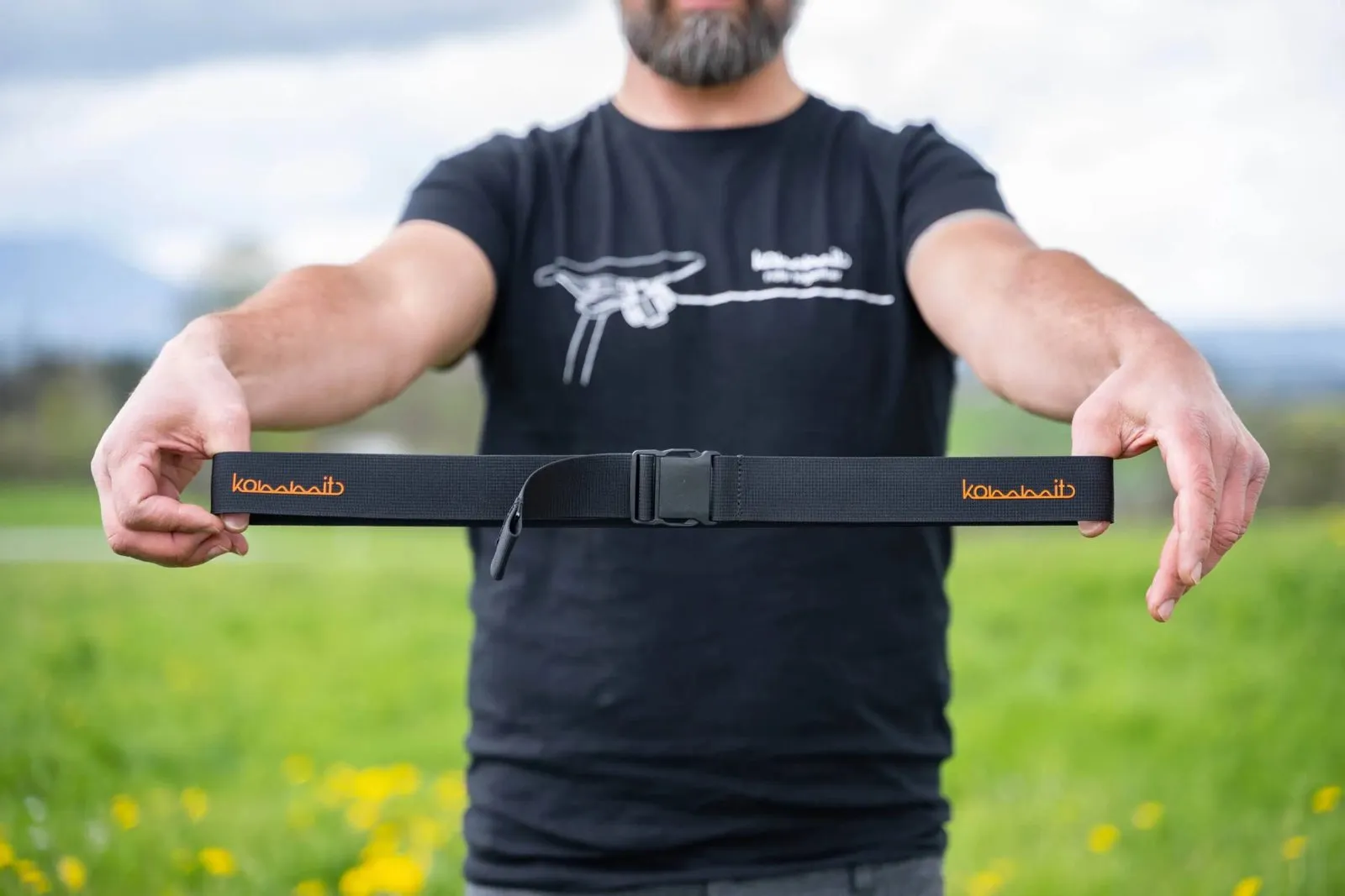 A person is holding a black outdoor belt with orange "kommit" lettering against a green background.