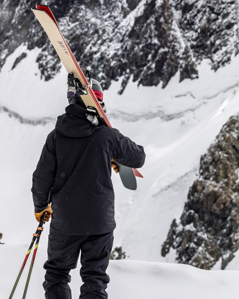Skieur devant une montagne Personne avec skis et bâtons devant une montagne enneigée, portant une veste noire S-1 Gore Tex.
