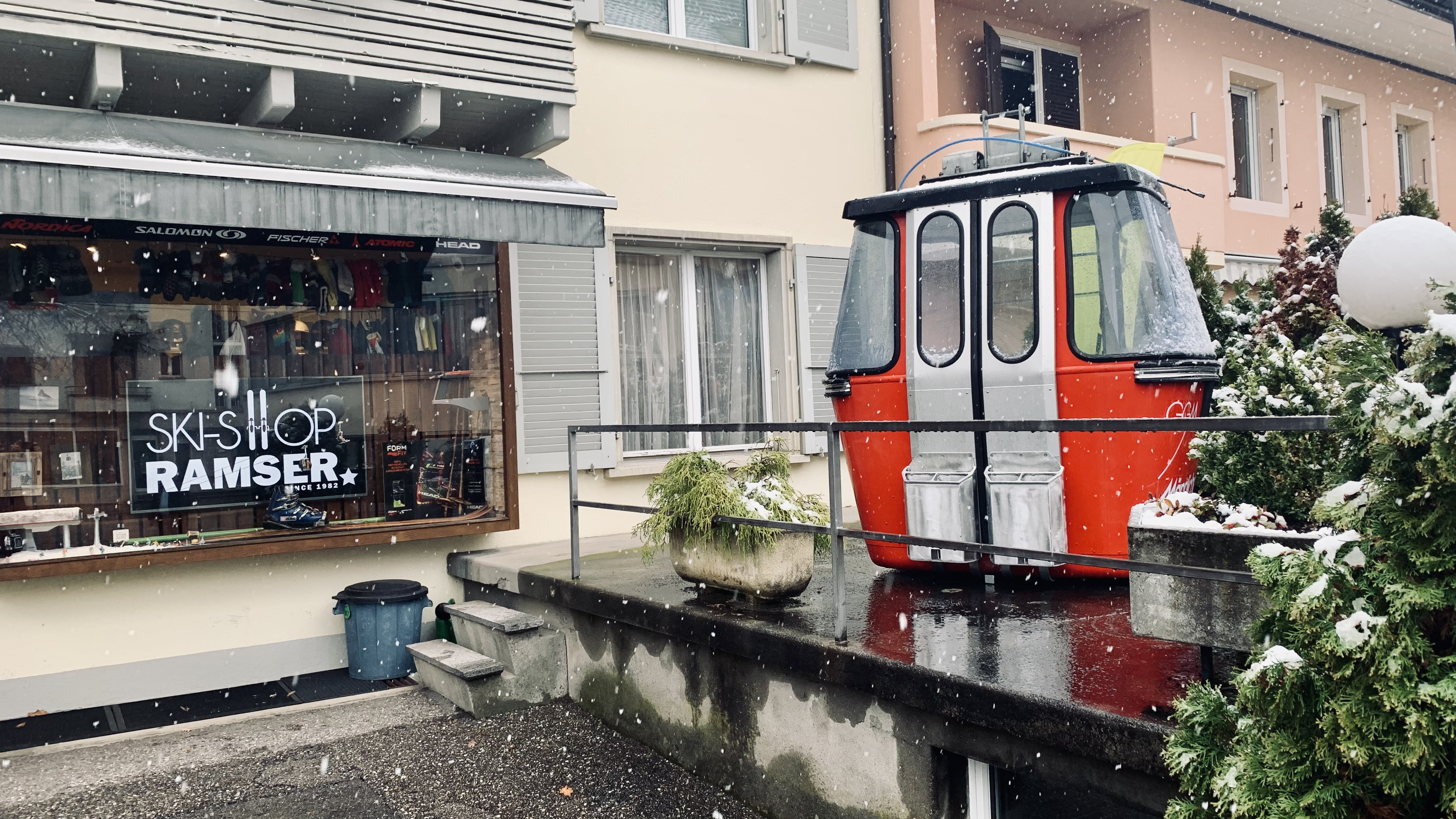 ski-shop.ch startet in die Neue Ski-Saison 21/22 Winterliches Stadtbild: Ski-Shop mit Gondel davor. Schnee fällt auf das rote und silberne Gondelmodell, die Fassade und die Pflanzen.