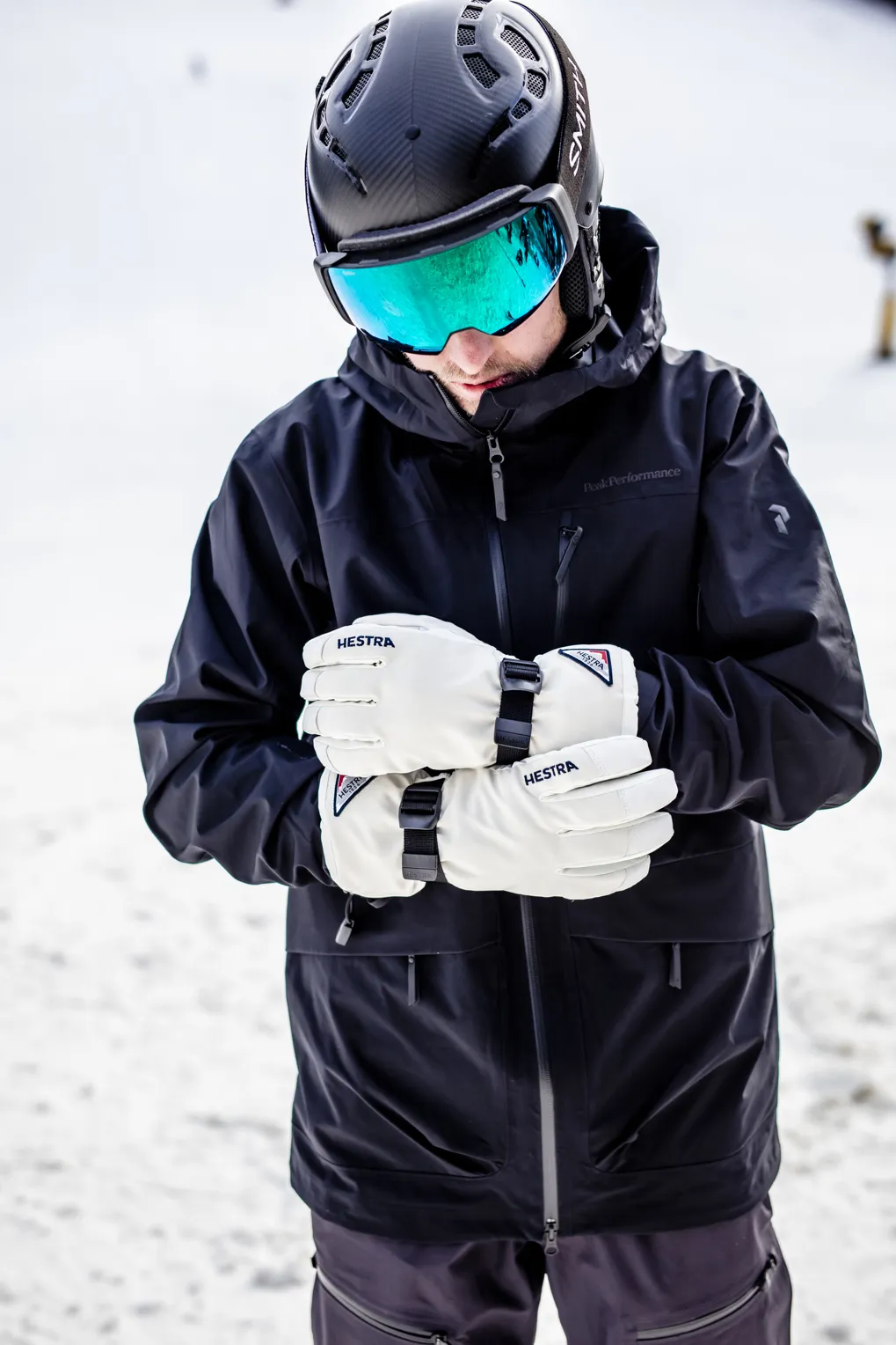 Mann mit Skihelm, Brille und Handschuhen vor verschneitem Hintergrund. Er trägt eine schwarze Jacke.