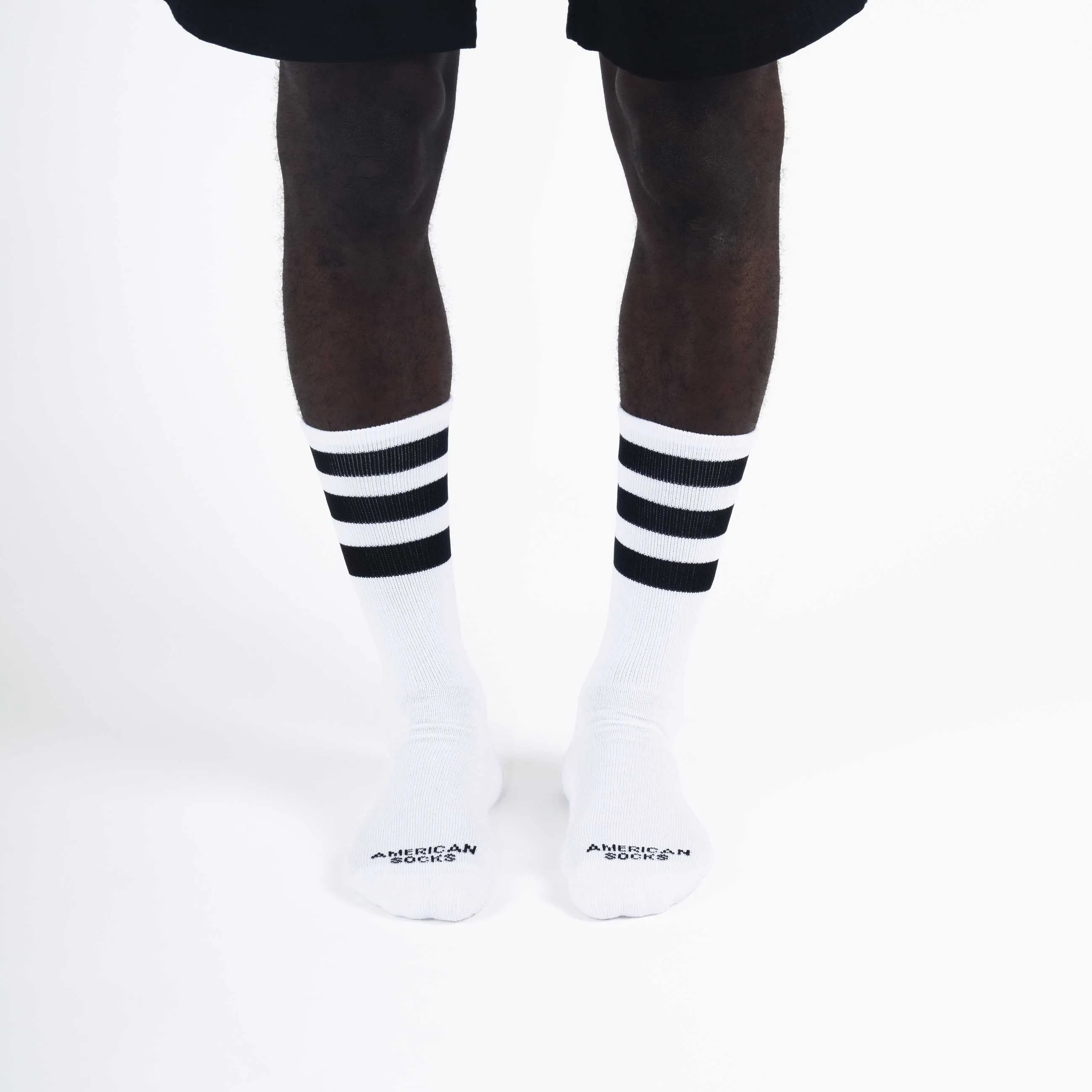 A man wears white, ribbed "Old School II - Mid High" socks with three black stripes, brand "American Socks".