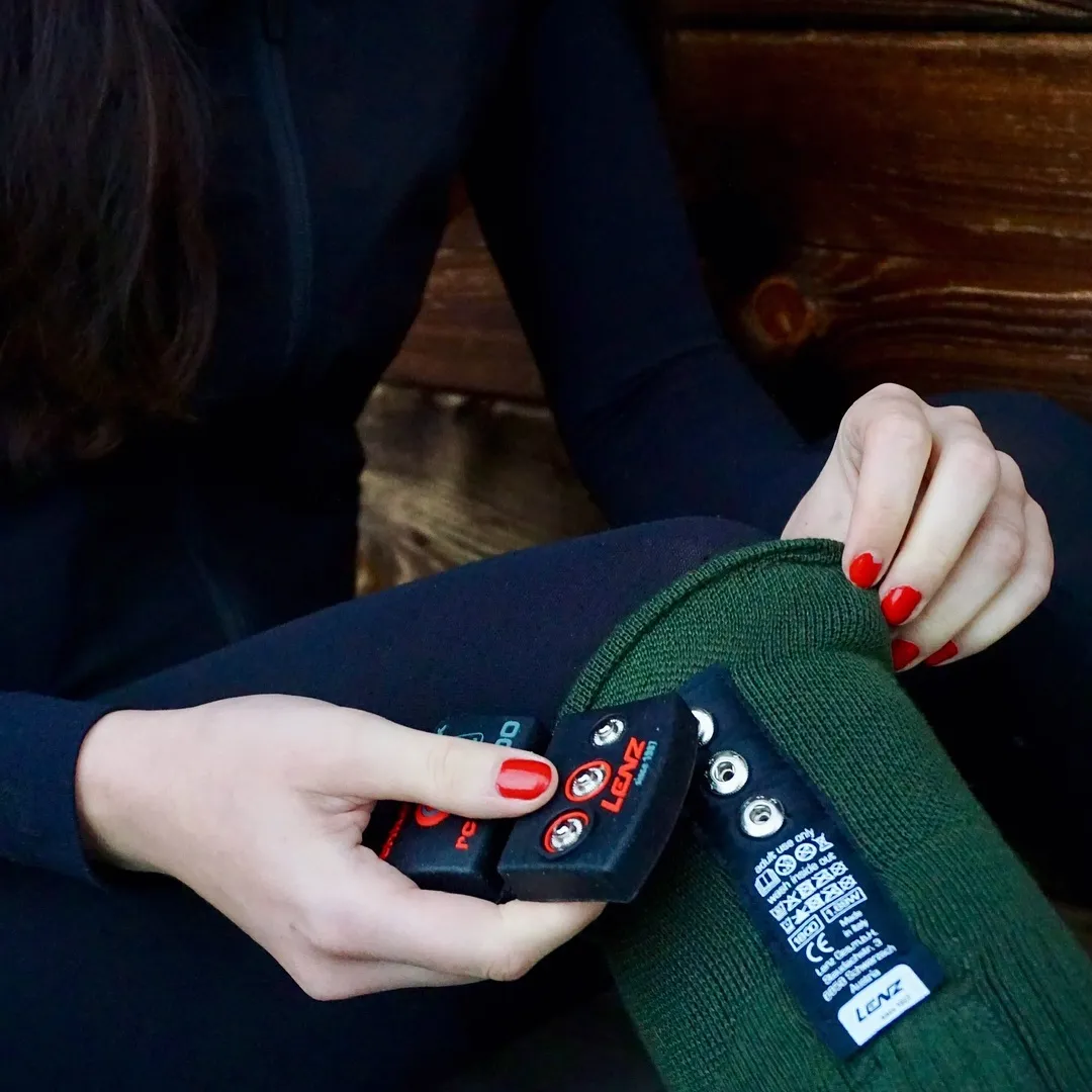 Una persona è seduta con leggings neri, tiene in mano batterie nere con bottoni rossi e un calzino verde con il logo Lenz Socken.