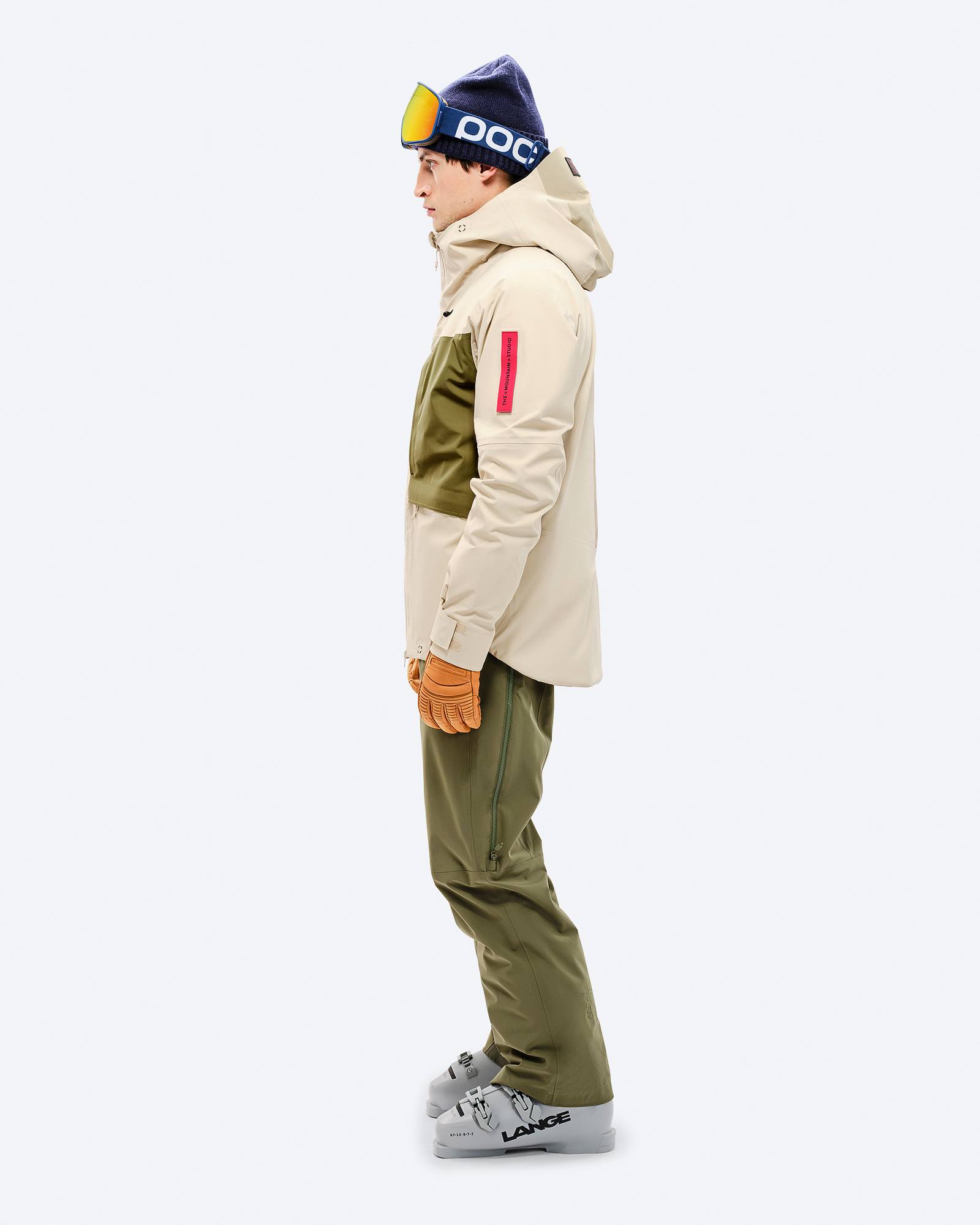 Ski-Outfit für Herren Mann in Ski-Kleidung, seitlich. Beige 3L Hybrid Shell Jacke, grüne Hose und Skischuhe. Blaue Mütze mit Skibrille.