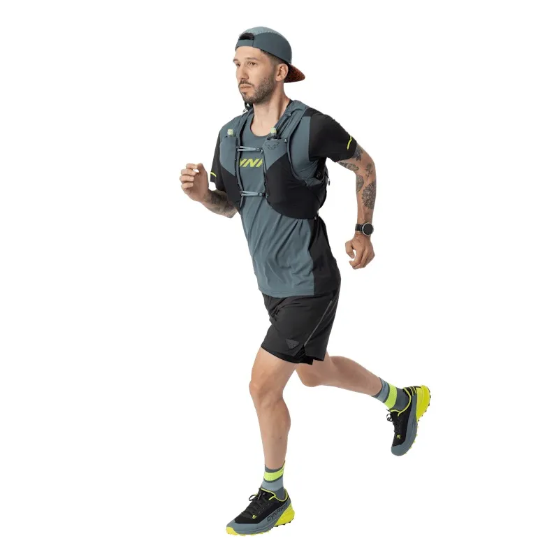 A man with a beard, a cap, and a running vest is running in front of a white background. He is wearing a gray running shirt, black shorts, athletic shoes, and a watch. He has a tattoo on his left arm.