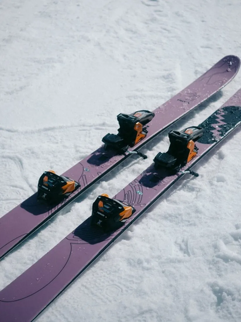 Due sci viola con attacchi arancioni e neri giacciono nella neve. Gli sci hanno un motivo grafico.