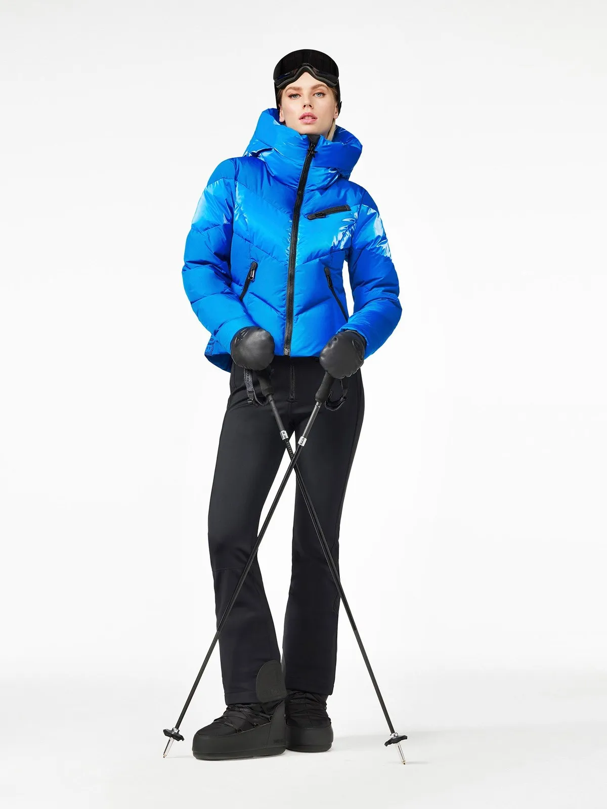 Frau in blauer Moraine Skijacke, schwarzer Hose, Skibrille und Handschuhen hält Skistöcke.