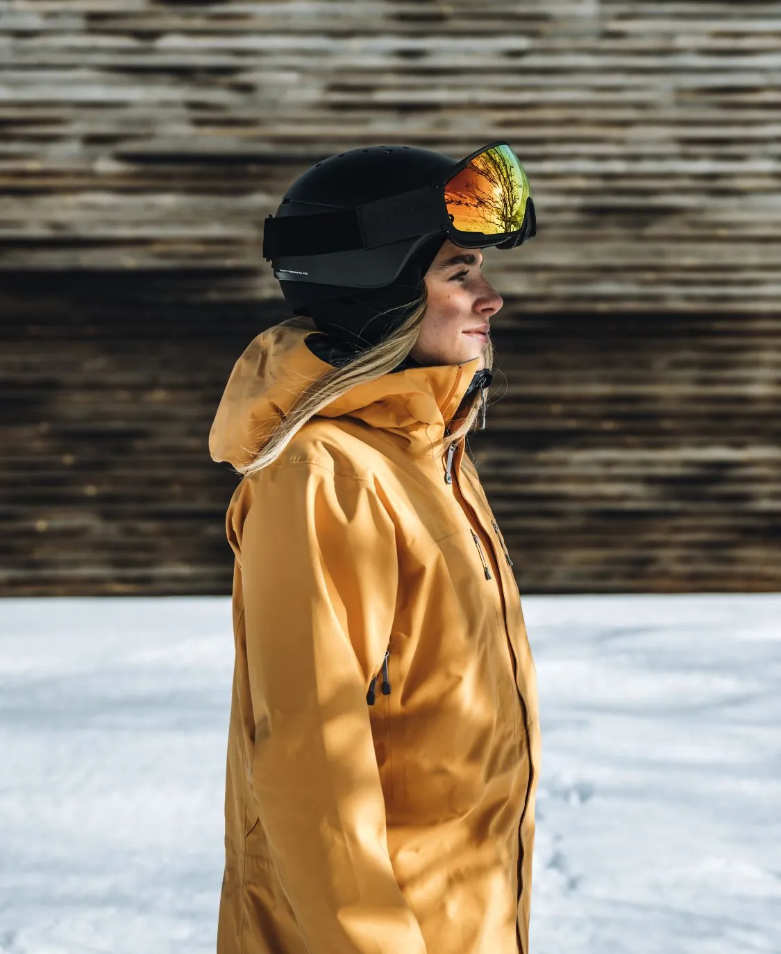 Vista di profilo di una persona che indossa un casco da sci POC Meninx nero e una giacca gialla sulla neve. Gli occhiali da sci riflettono gli alberi.
