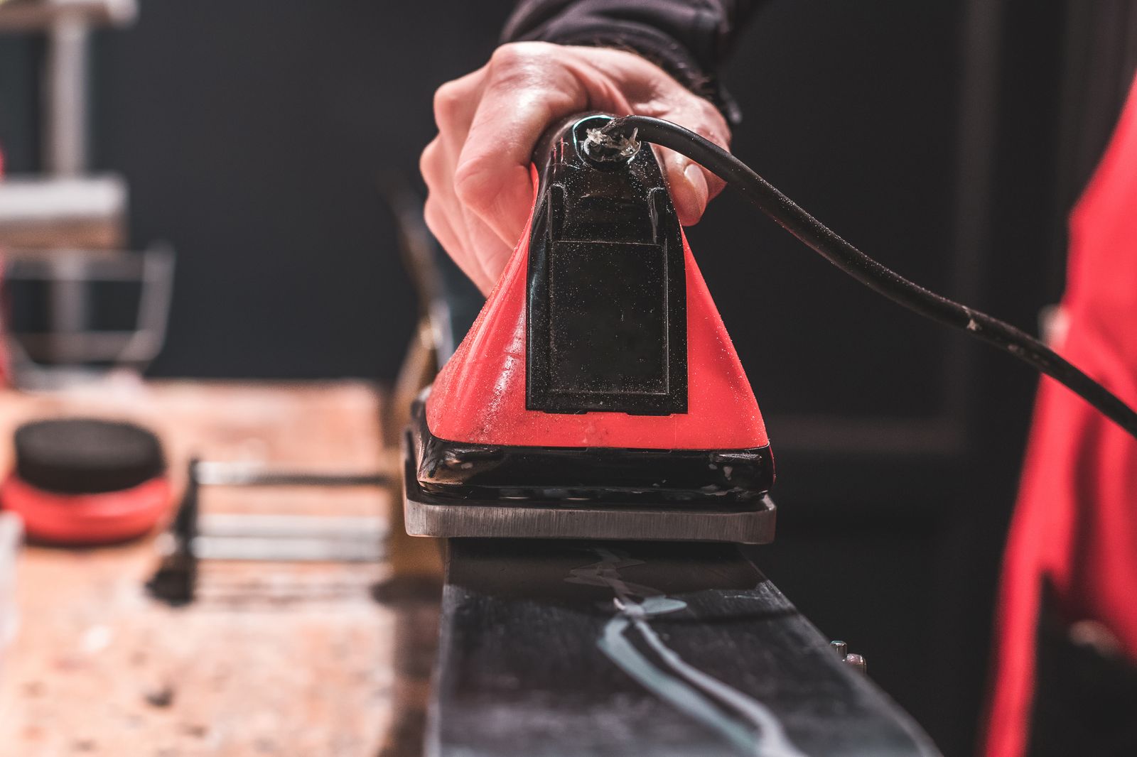 Une main tient un fer à repasser rouge et noir au-dessus de la surface d'un ski. Le fer à repasser glisse sur le ski pour appliquer le fart.