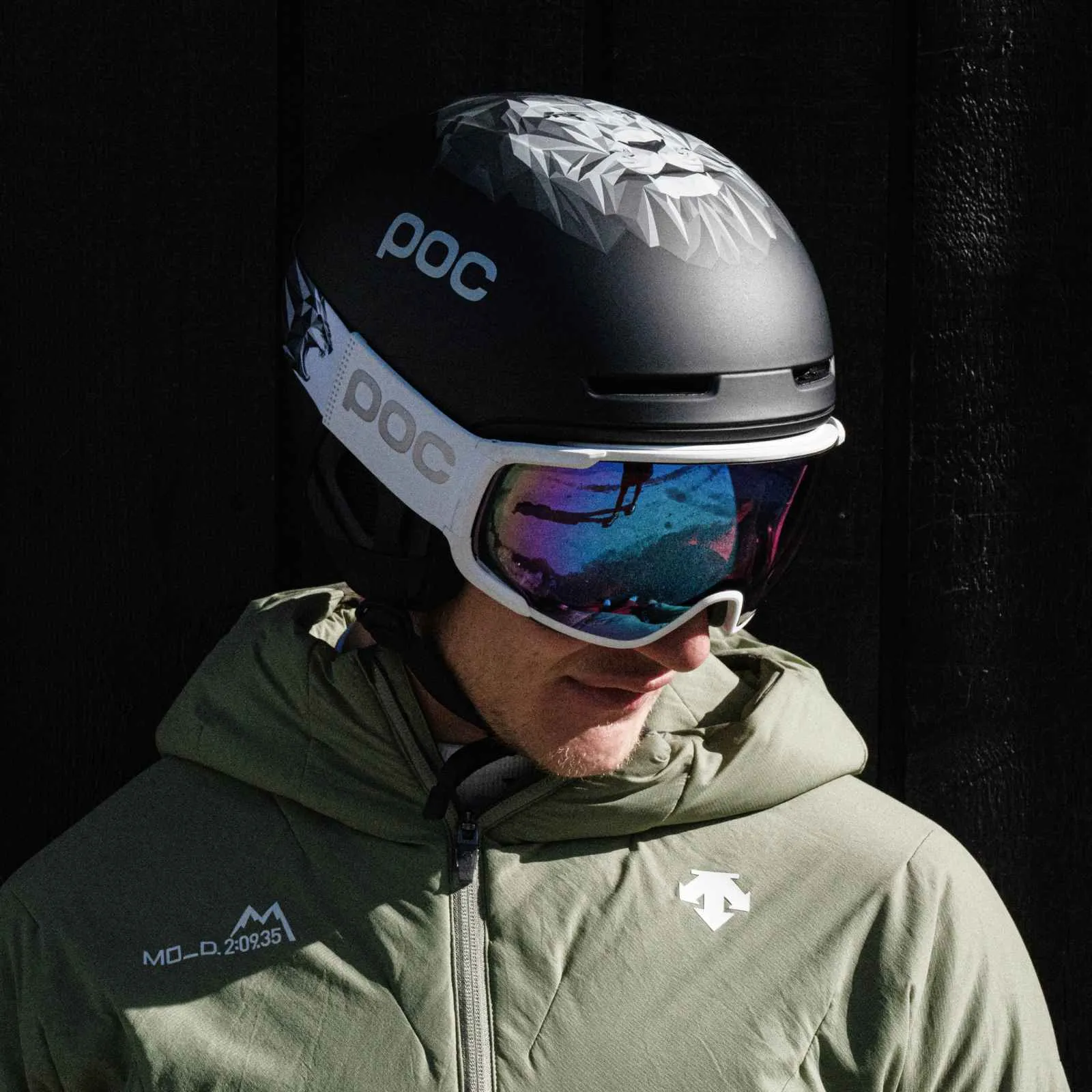 Homme avec casque de ski POC, motif Lwen, et lunettes de ski. Réflexion dans les lunettes. Veste verte.