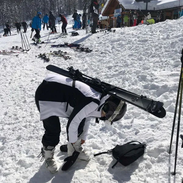 Skier bends over to fasten ski boots, skis with ski rack on her back. Snowy landscape.