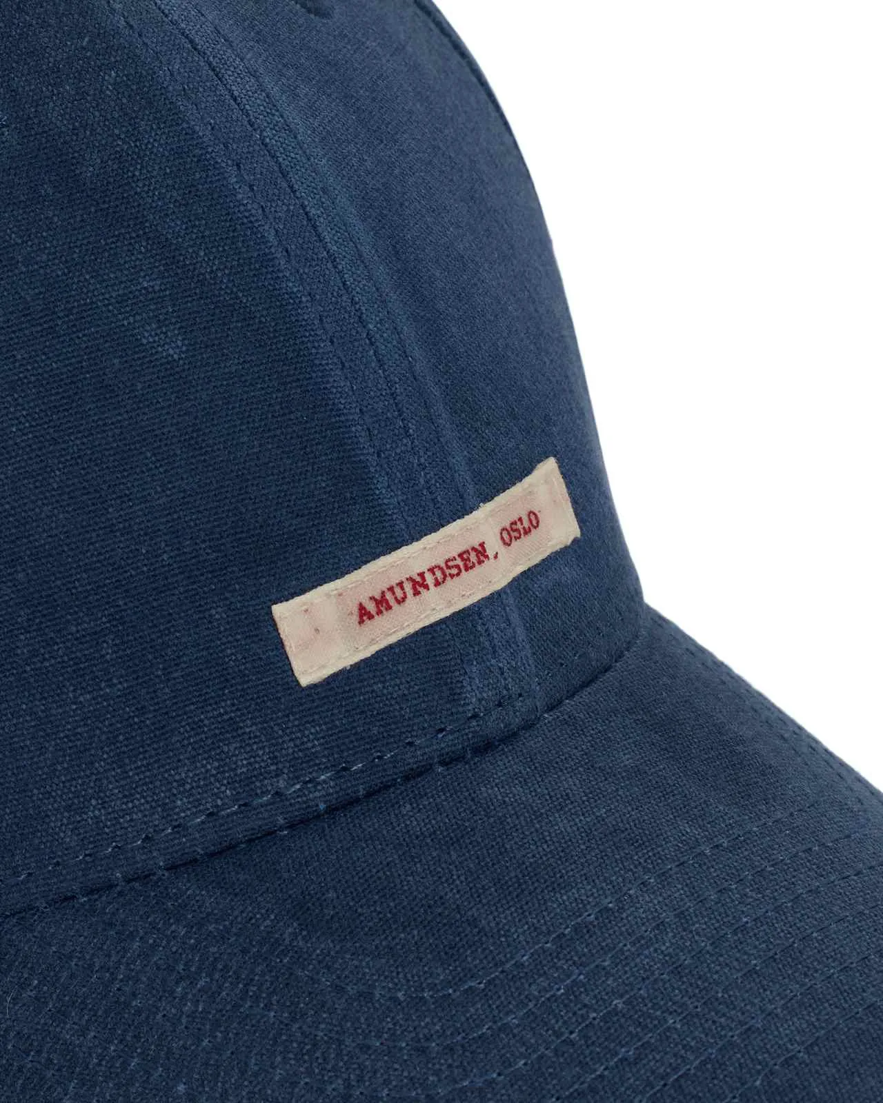 Eine blaue Baseballkappe ist aus einem groben Stoff gefertigt. Ein rechteckiges cremefarbenes Etikett ist an der Seite der Kappe befestigt, mit dem Text „AMUNDSEN, OSLO“ in roter Schrift. Nähte akzentuieren den Schirm und die Seiten der Kappe.