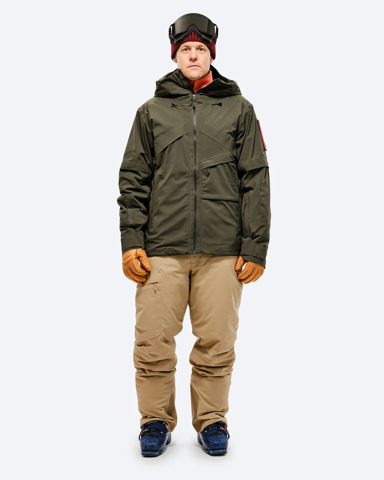 Person in ski clothing: olive green jacket, sand-colored pants and ski boots against a white background.