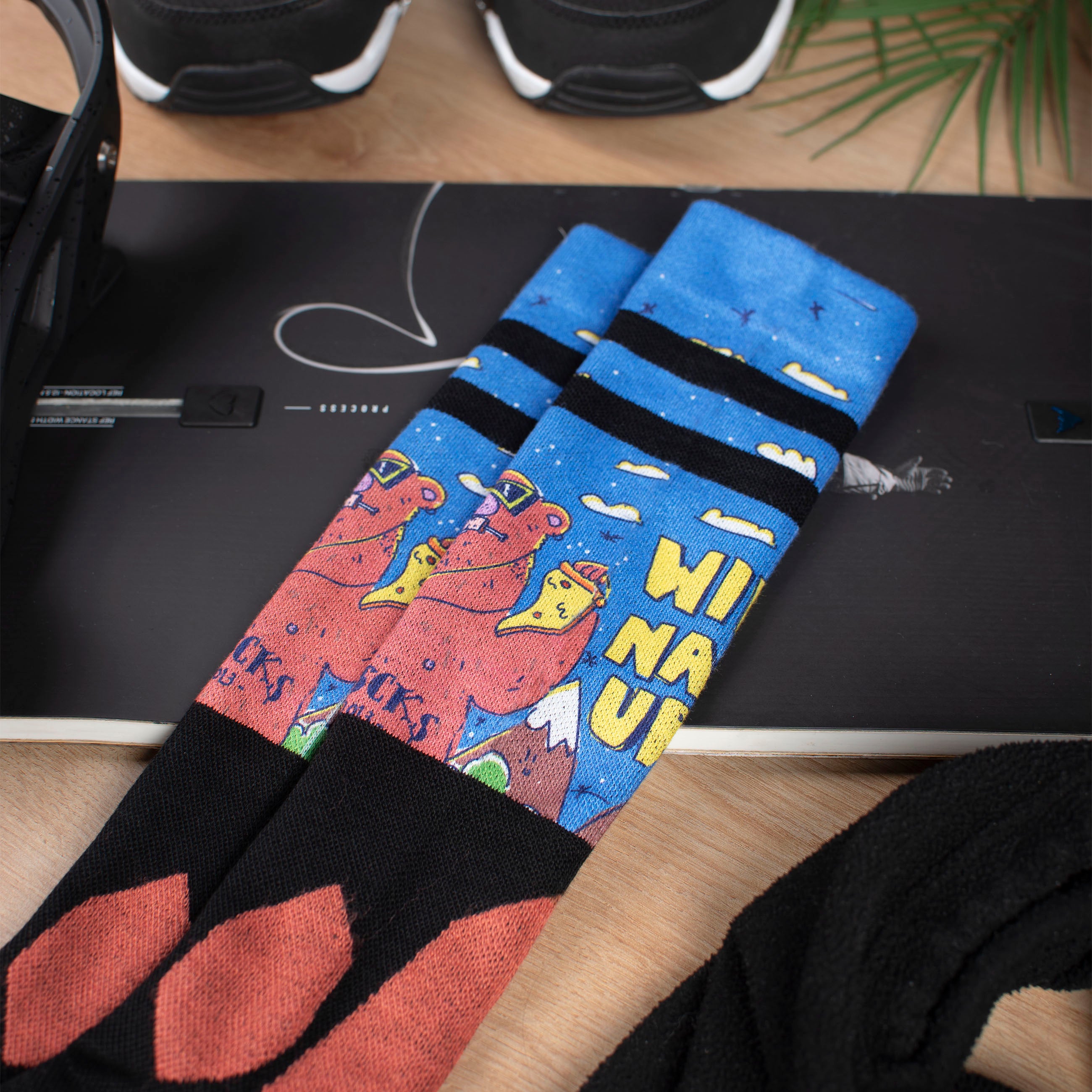 Bärenmotiv Schneesocken Snow Socks mit Bärenmotiv liegen auf einem Snowboard, Boots und Grün im Hintergrund.