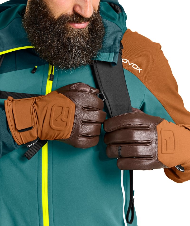 Nahaufnahme: Mann mit braunen Ortovox Alpine Pro Handschuhen, greift Rucksackträger, Jacke in Grün/Braun.