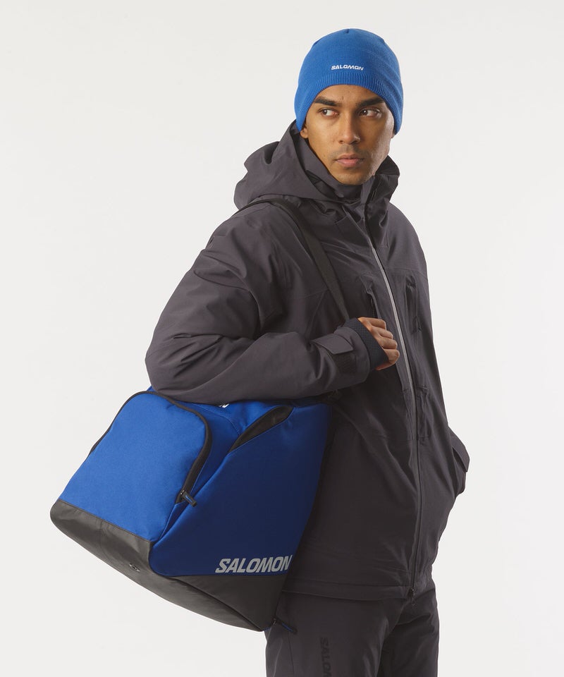 Homme avec une casquette bleue, portant Salomon sac de chaussures de ski sur l'épaule. Le sac est bleu et noir avec le logo Salomon.