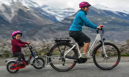 Frau und Kind mit FollowMe Tandemkupplung auf Fahrrädern vor Berglandschaft.