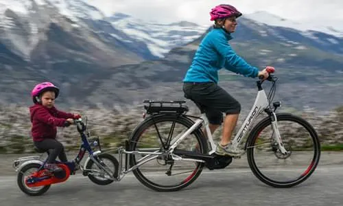 Woman and child with FollowMe tandem coupling on bicycles in front of a mountain landscape.