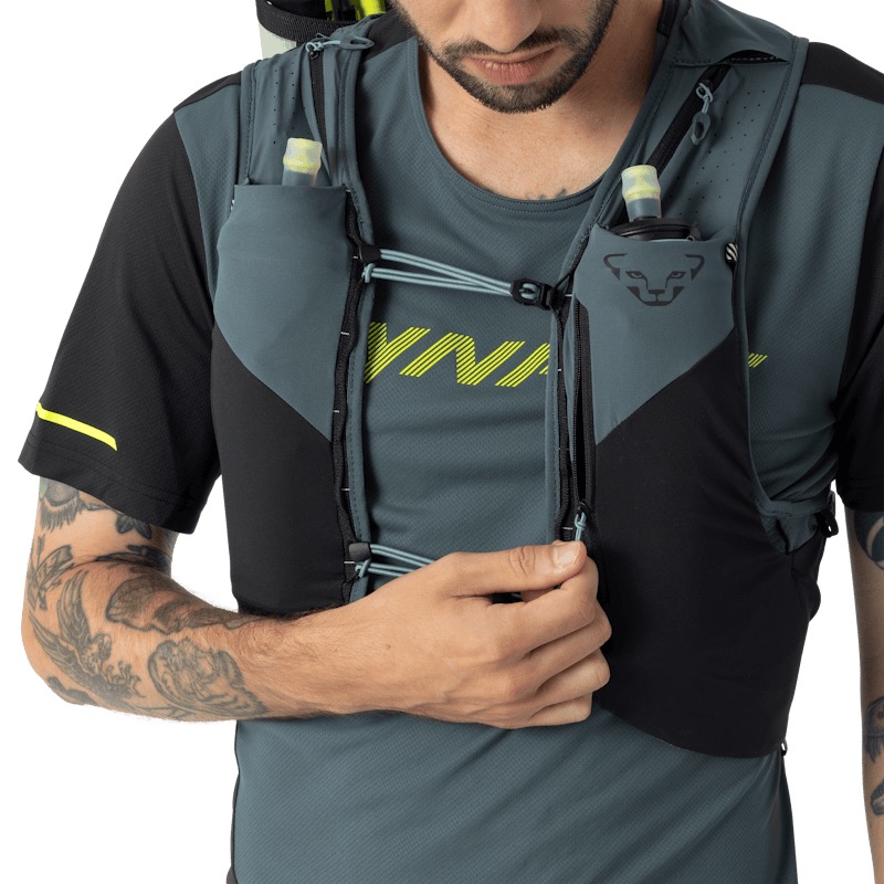 Un uomo con la barba indossa un gilet da corsa con borracce nelle tasche. Apre la cerniera del gilet. Il gilet è grigio e nero. Indossa una maglietta da corsa grigia con una stampa gialla e maniche corte nere. Ha un tatuaggio sul braccio.