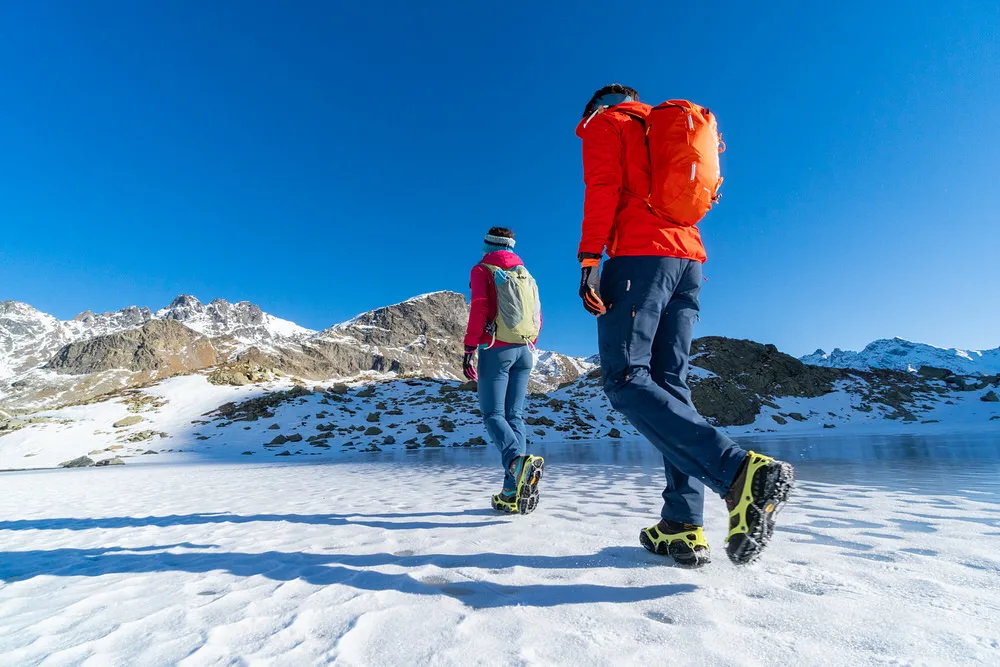 Due persone con artigli da scarpe veloci che camminano sulla neve sullo sfondo di una montagna.
