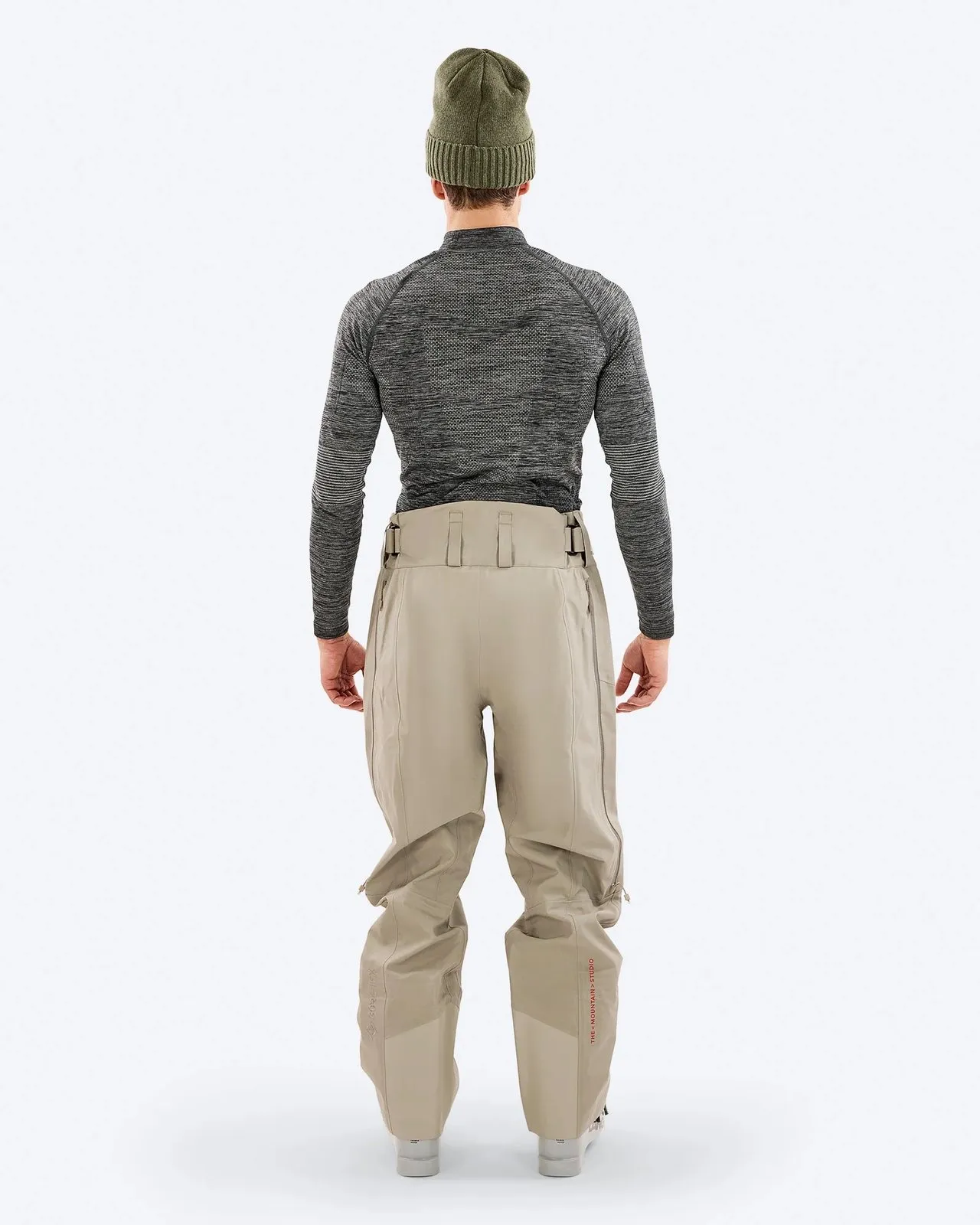 Rückansicht einer Person mit olivgrüner Mütze, graumeliertem Langarmshirt und beigen Skihosen vor weißem Hintergrund.