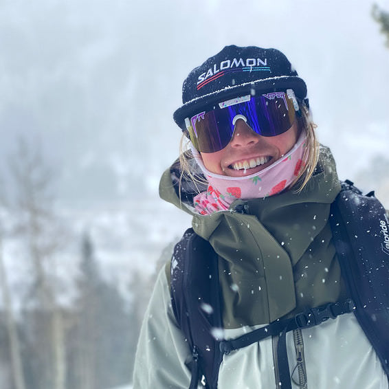 Frau im Schnee Frau im Schnee mit Pit Viper Jetski Elliptical Sonnenbrille, Mütze und Erdbeer-Tuch lächelt.