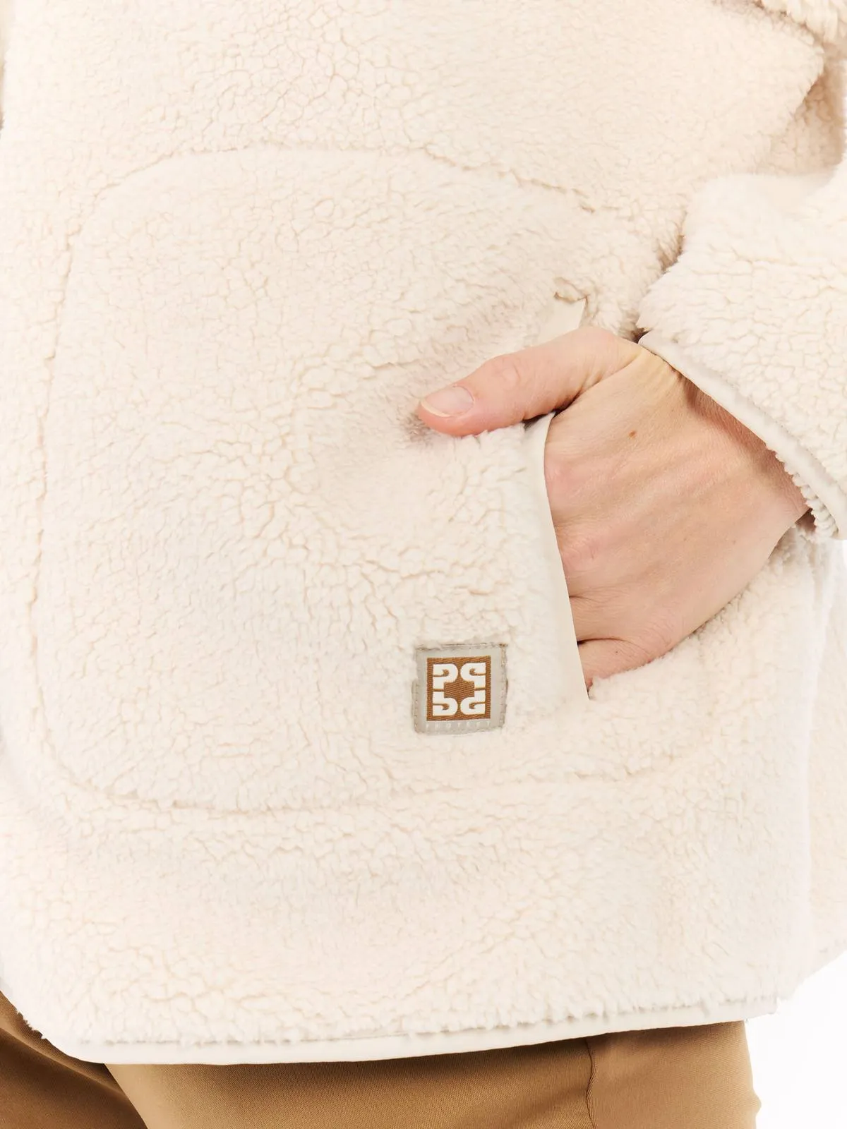 Primo piano di una giacca in pile beige con una mano nella tasca e un piccolo logo quadrato sul lato sinistro.