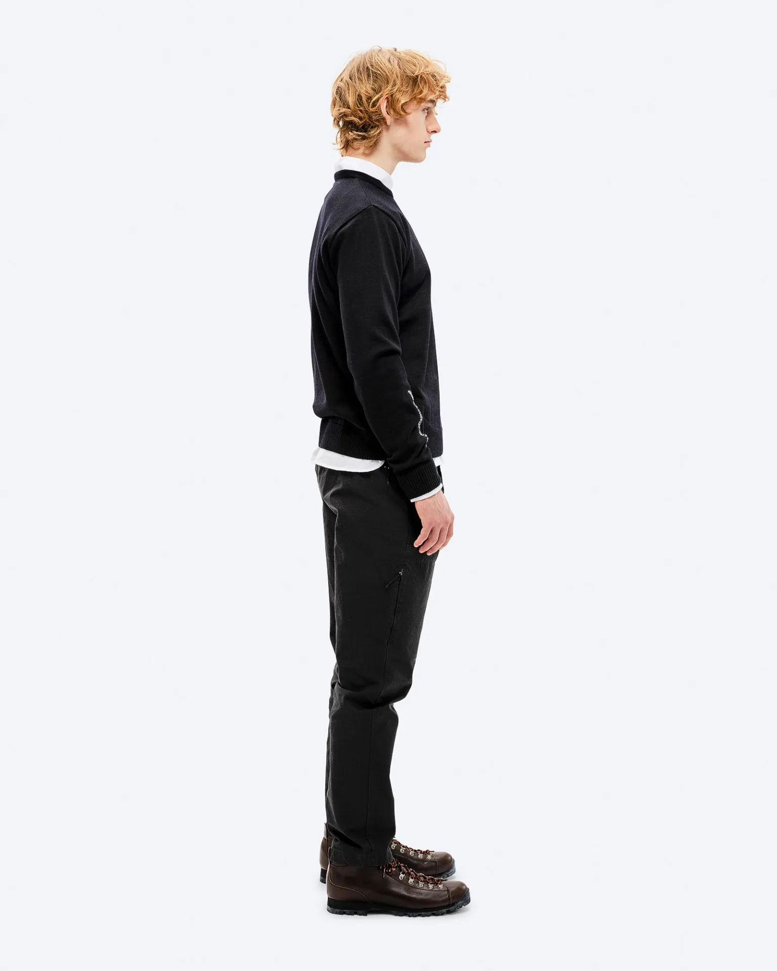 Ein Mann im Profil trägt einen schwarzen K-3 Ski Sweater aus Merinowolle, dazu dunkle Hosen und braune Wanderschuhe.