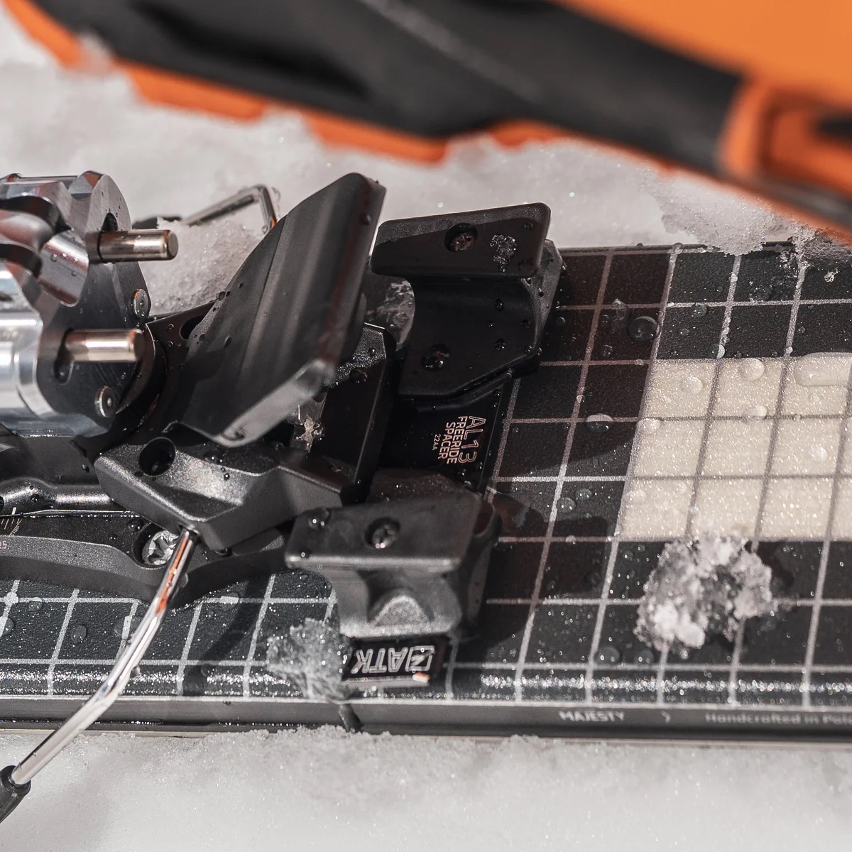 Nahaufnahme einer Skitourenbindung von ATK auf einem Ski mit einem schwarz-weißen Schachbrettmuster. Schnee liegt auf dem Ski.