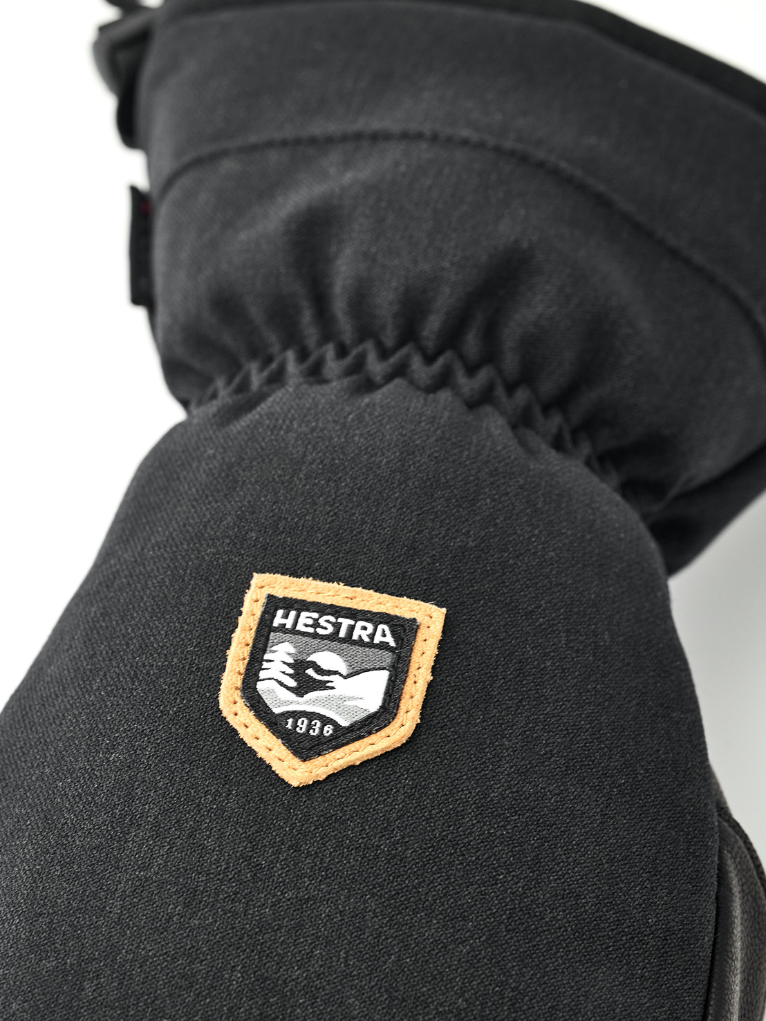 Black mitten with logo Close-up of a black mitten with the Hestra logo, an orange-bordered shield with a mountain motif and the founding year 1936.