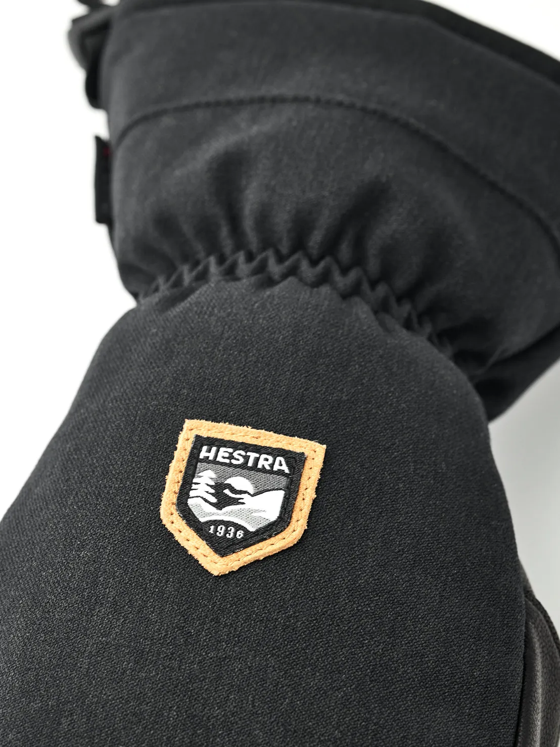 Détail d'une moufle noire avec le logo Hestra, une plaque orange bordée d'un motif de montagne et l'année de fondation 1936.