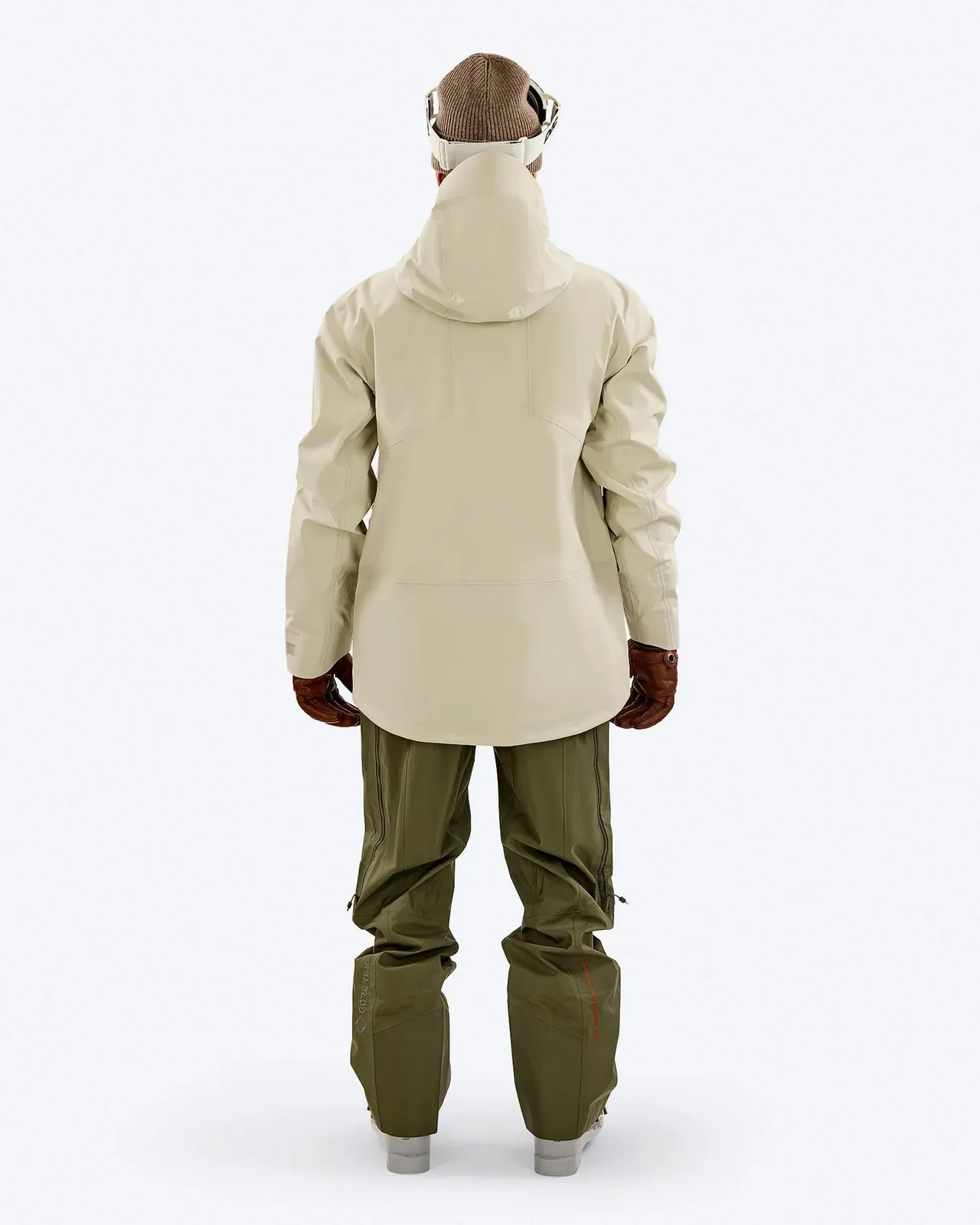 Rear view of a person in ski clothing: beige jacket, green pants, brown gloves, cap and ski goggles on a white background.